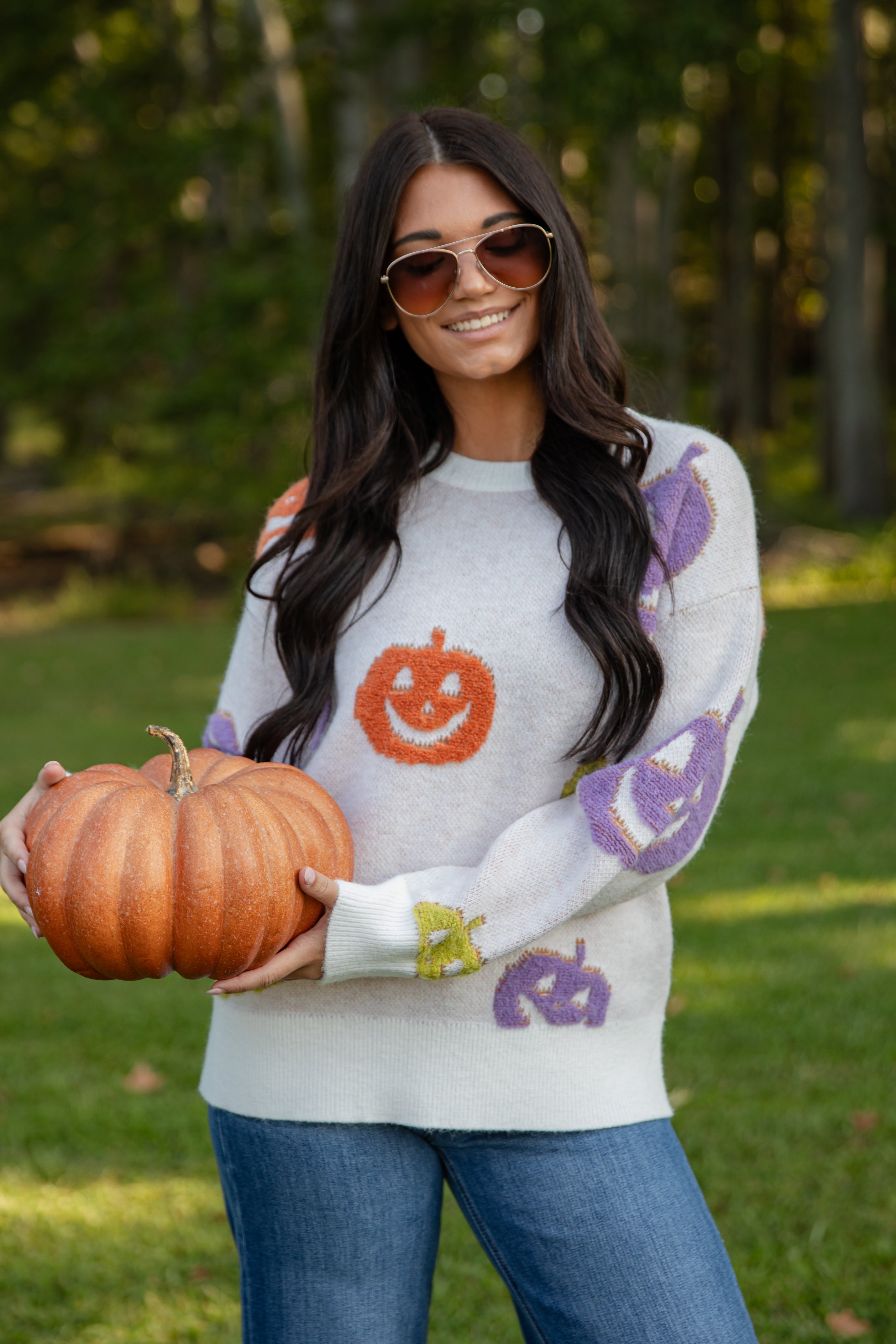 Woman wearing a Halloween-themed sweater holding a pumpkin in an outdoor setting.