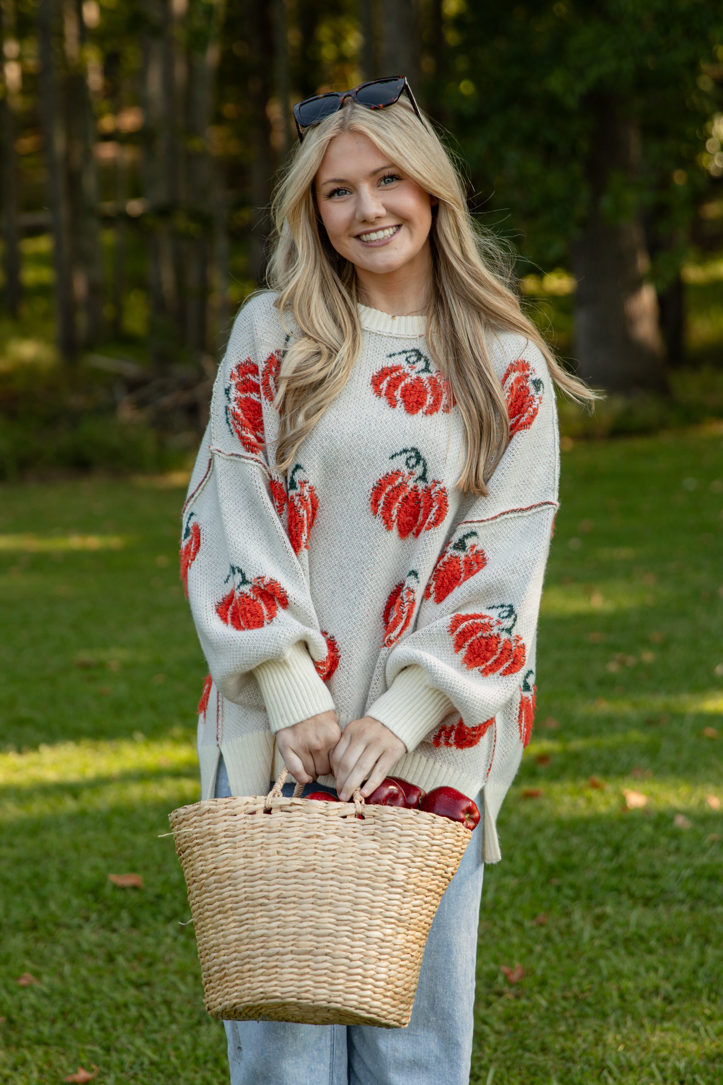 Woman wearing a white sweater with red patterns, holding a woven basket in a forest setting.