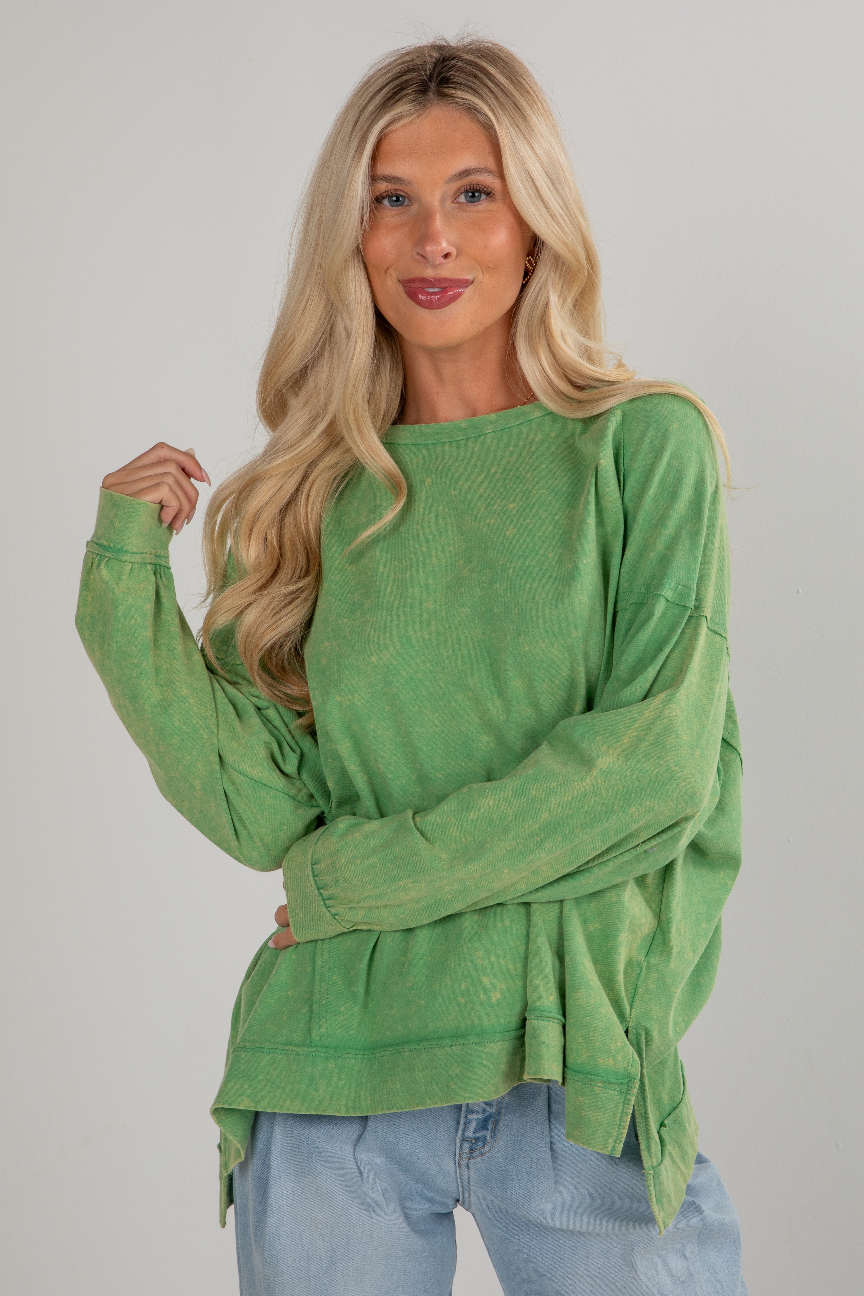 Woman wearing a green long-sleeve top and blue jeans against a gray background