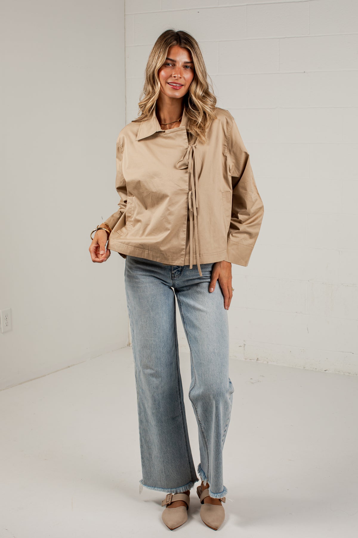 A woman modeling a light tan utility jacket with front tie details, paired with wide-leg light blue jeans and beige pointed-toe flats, standing against a white brick wall.