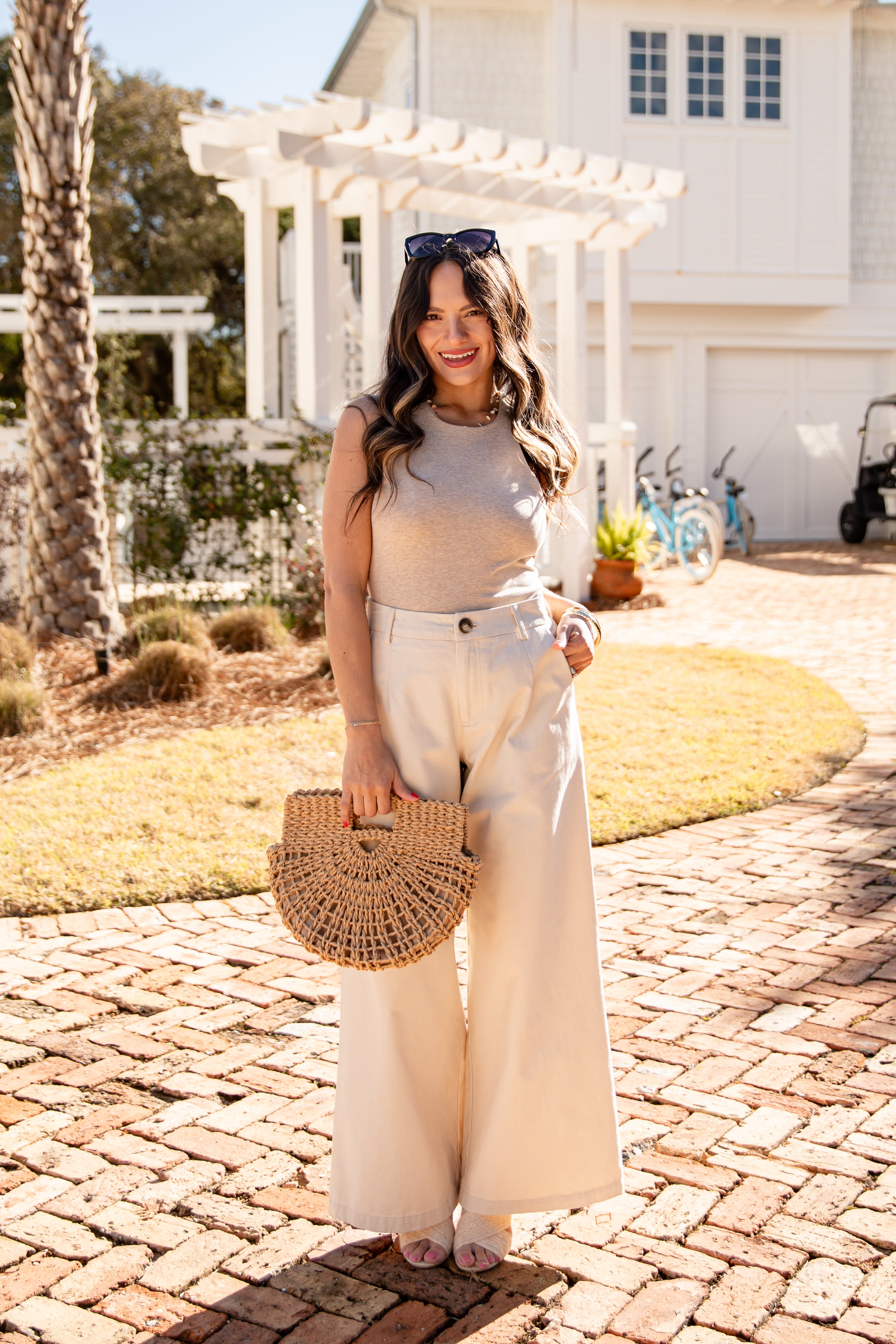 Woman standing on a brick path holding a woven bag, wearing a beige top and white pants, with a house and garden in the background.