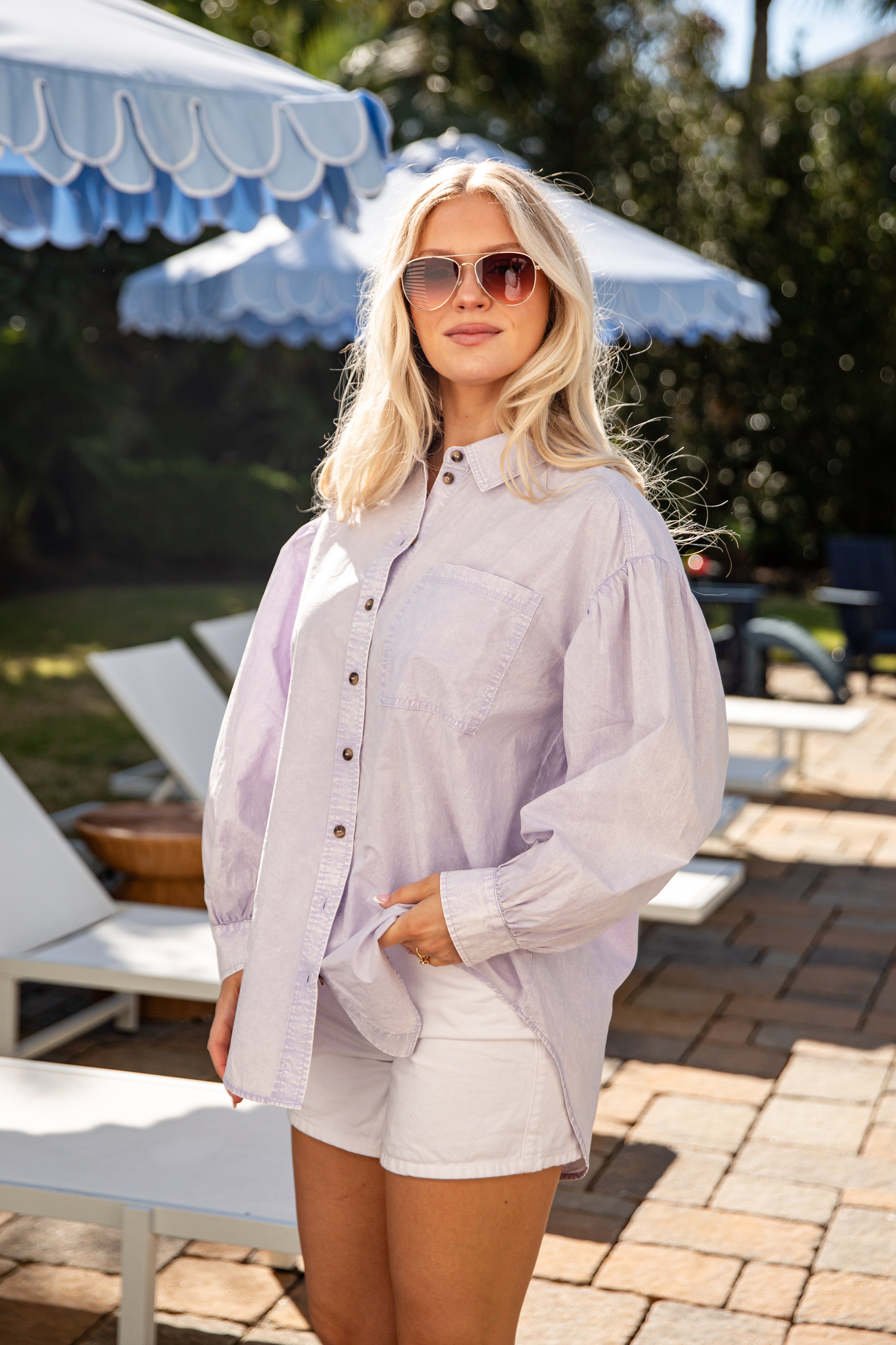 Woman in light purple shirt and white shorts standing outdoors with umbrellas in the background