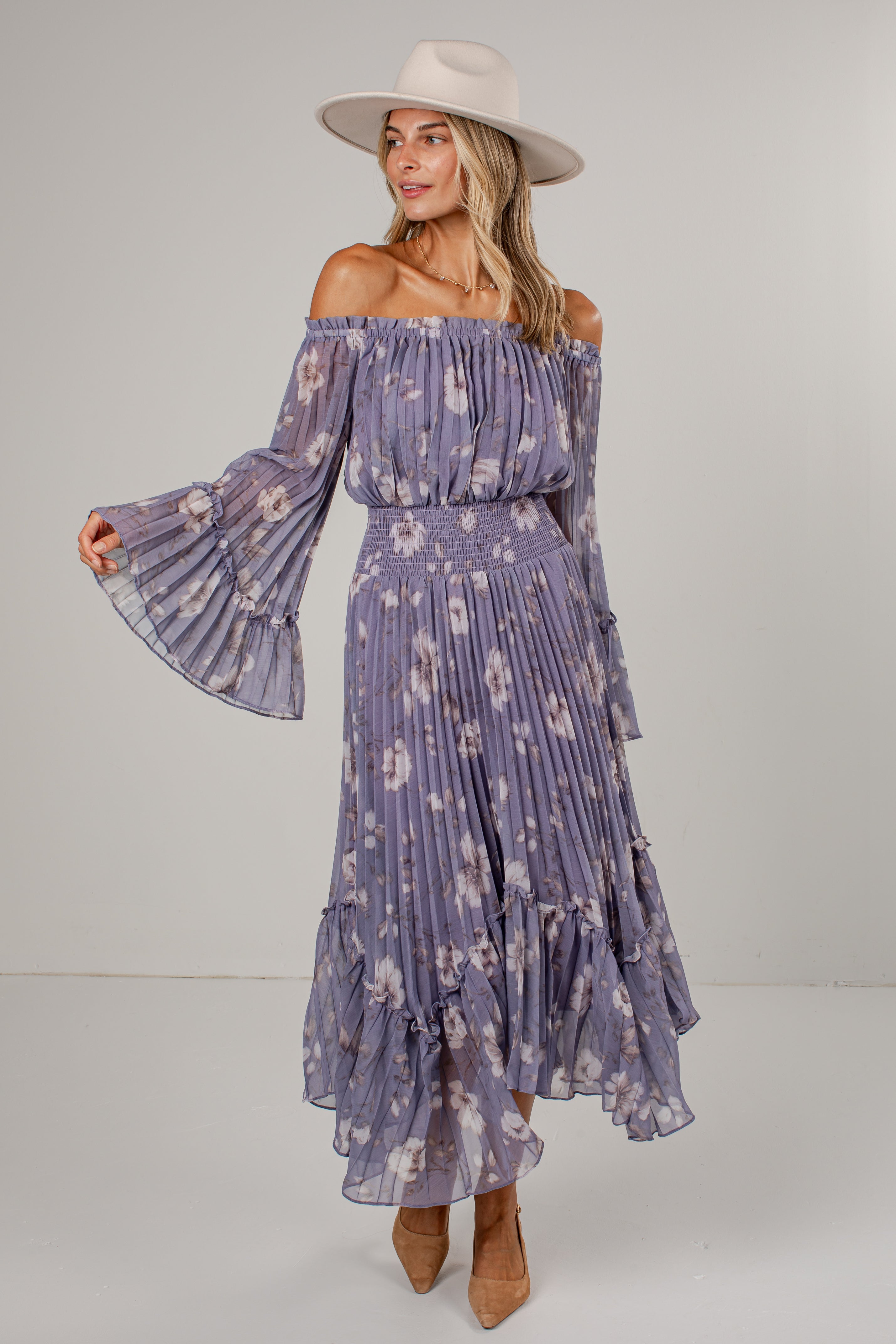 A woman modeling an off-the-shoulder midi dress with a purple and white floral print, featuring long bell sleeves, a smocked waist, and a tiered ruffle hem. She is also wearing a beige wide-brim hat and tan ankle boots, set against a plain light gray studio background.