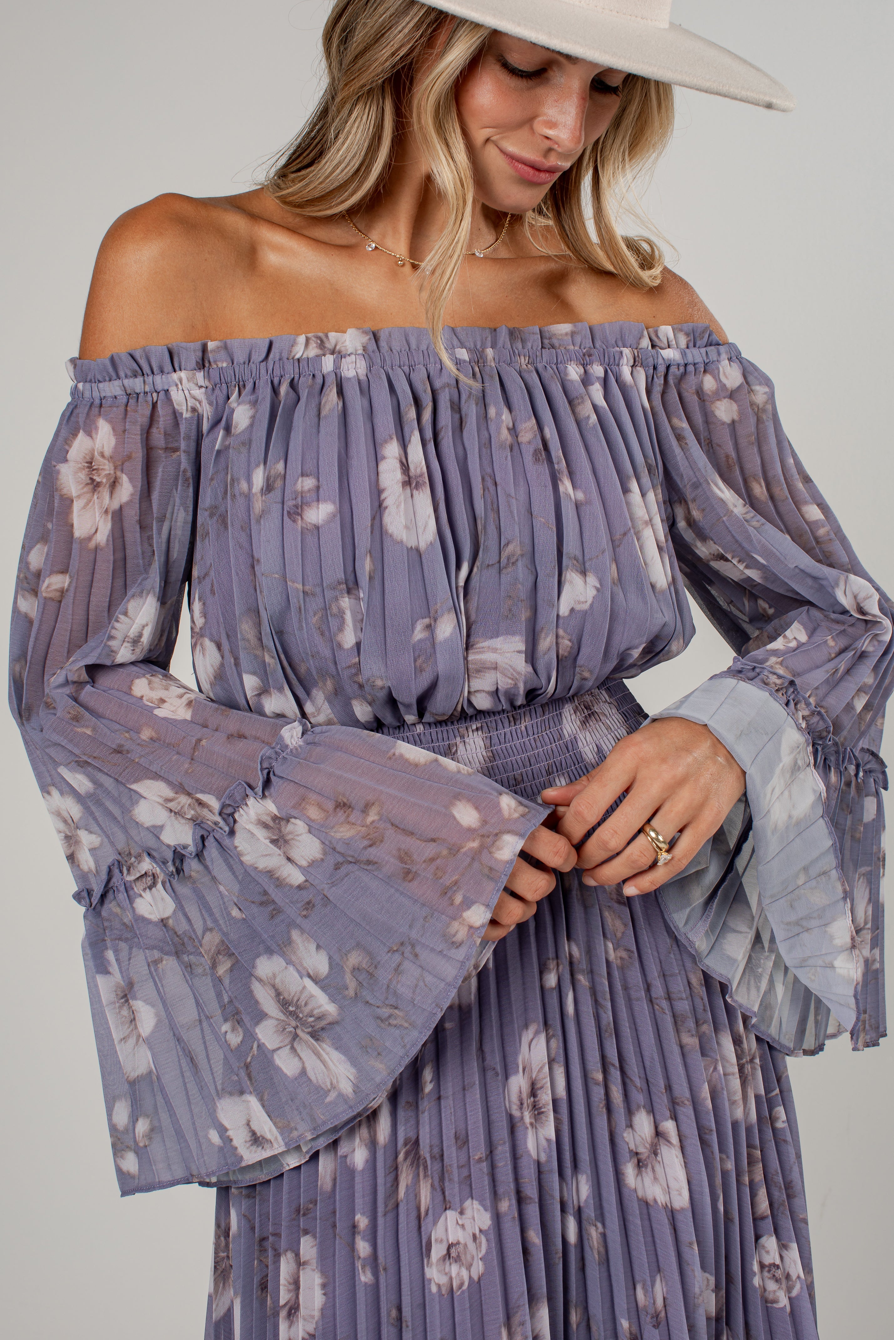 A woman wearing a lavender off-the-shoulder long-sleeve maxi dress with a white floral print, pleated fabric, and a matching pleated belt. She is also wearing a wide-brimmed cream hat, photographed against a neutral light gray background.