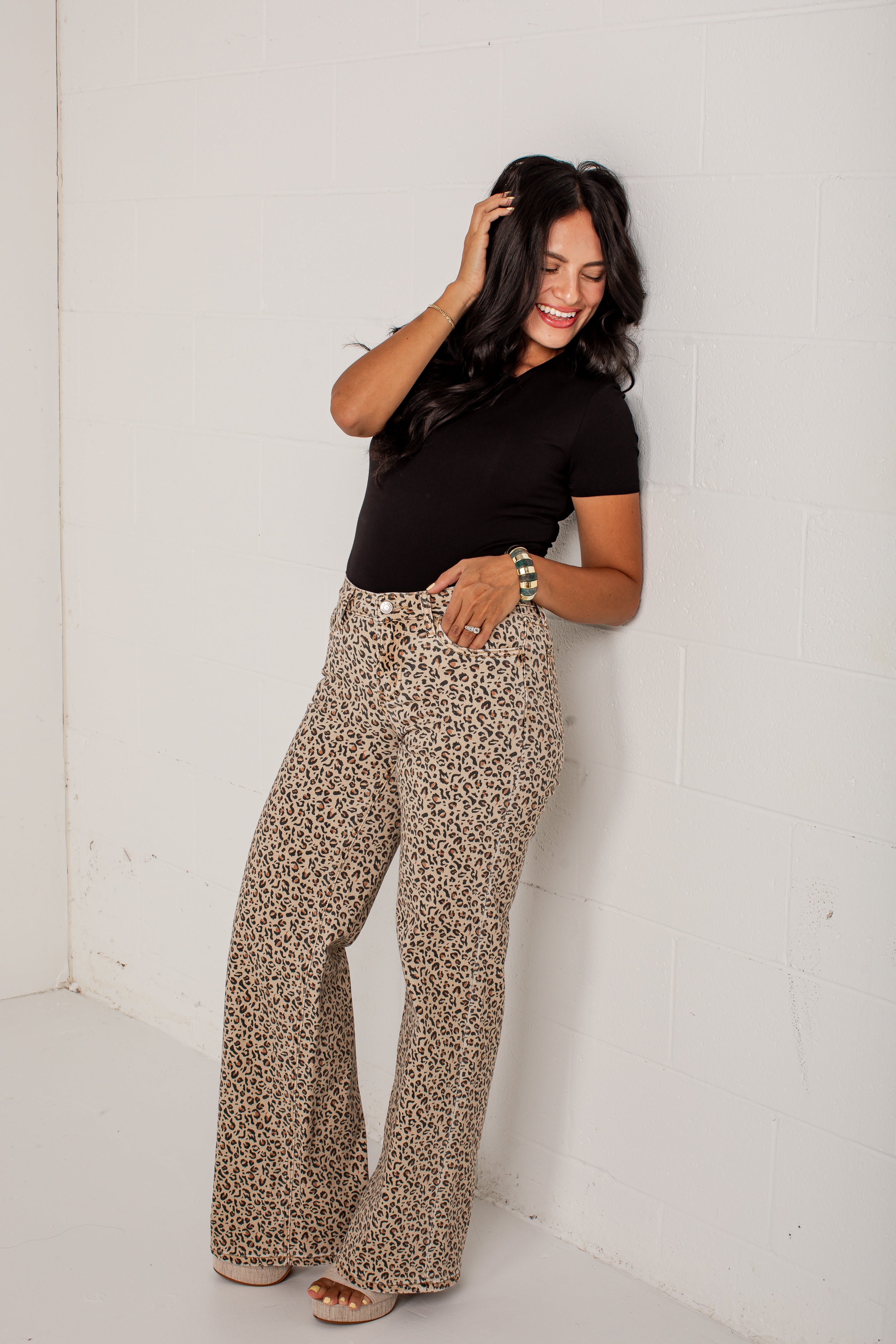 A woman posing in a white room corner wearing leopard print wide-leg pants and a black short-sleeve top.