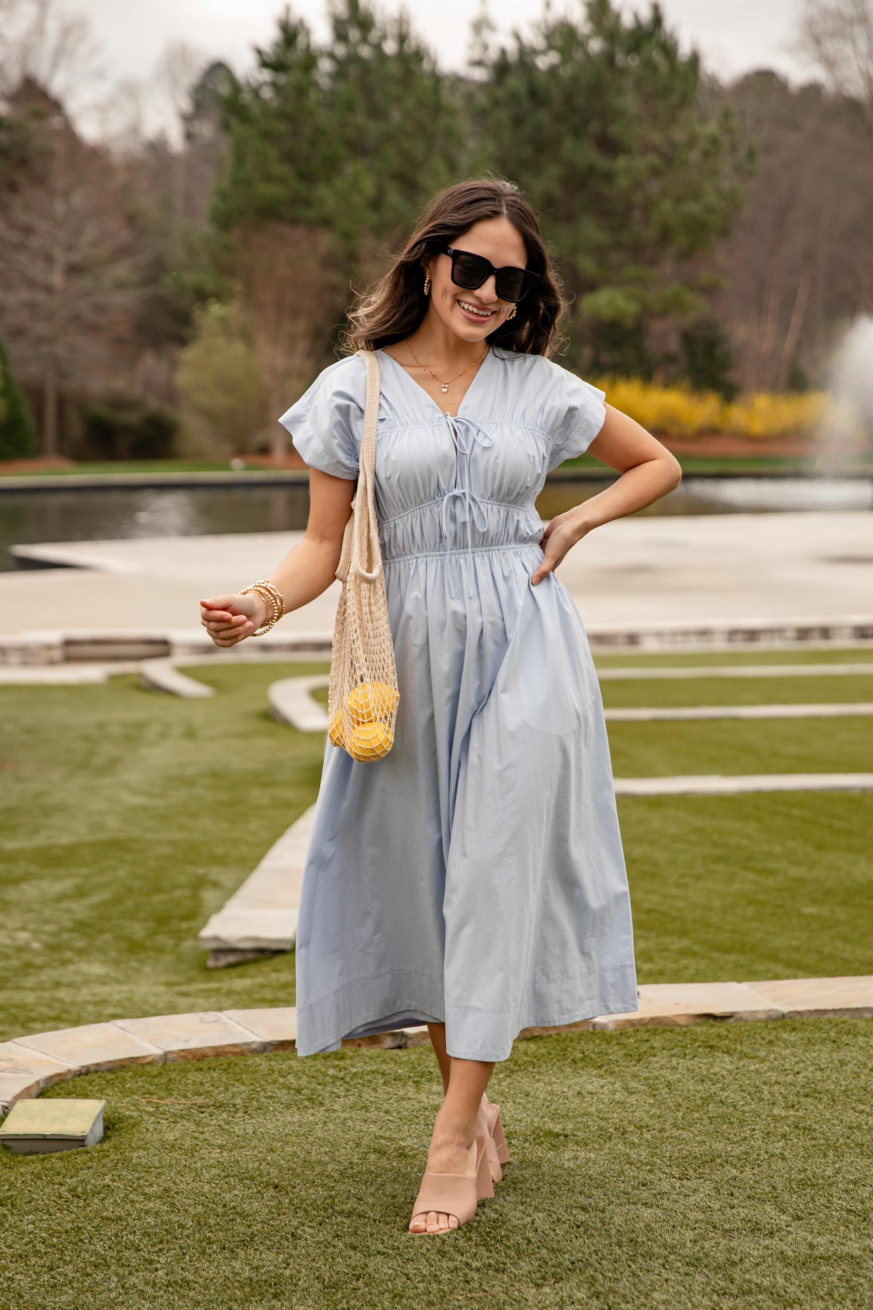 Woman in a light blue dress standing outdoors with a fountain and greenery in the background