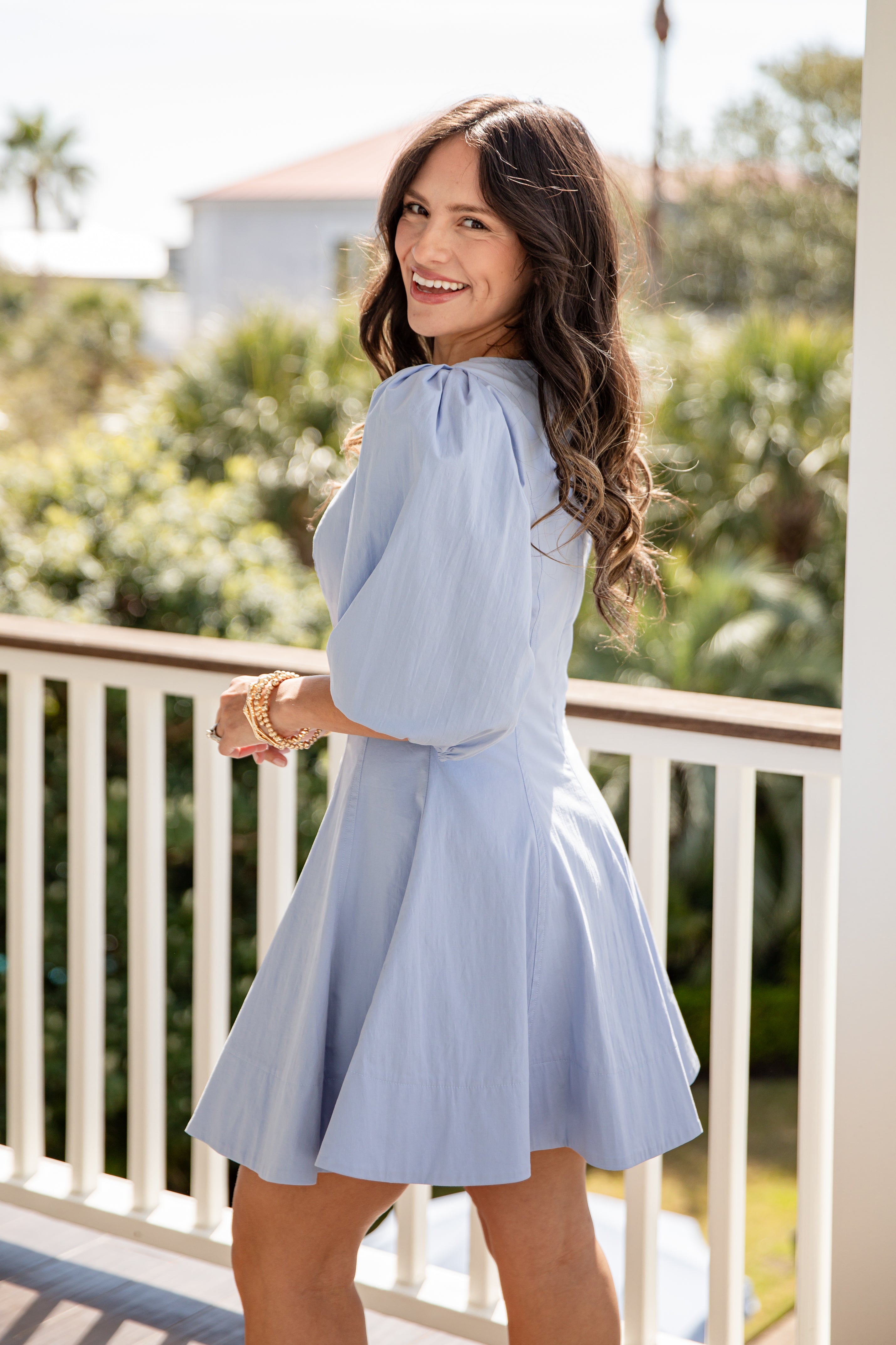 Woman in a light blue dress standing on a balcony with greenery in the background