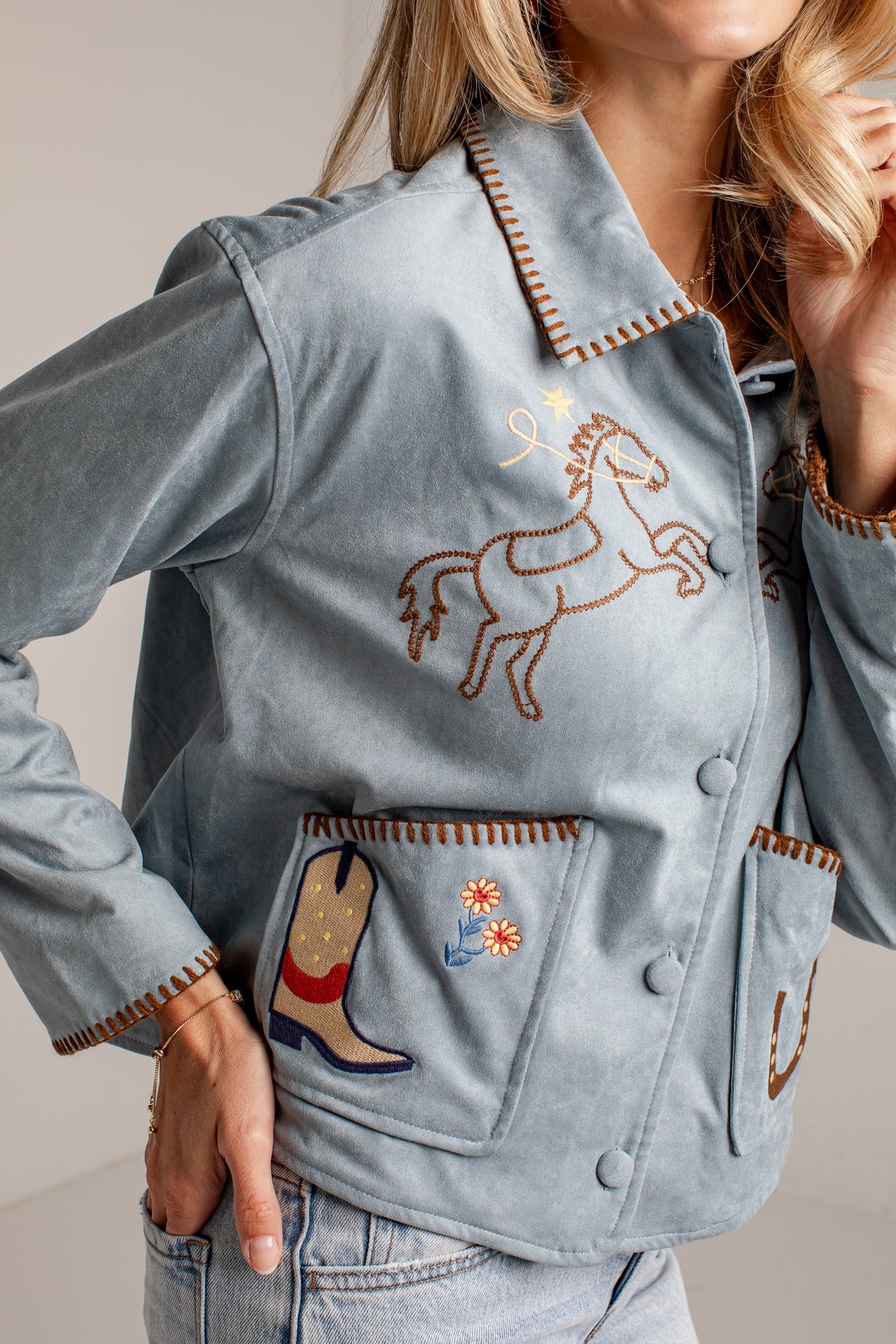 A close-up of a woman wearing a light blue suede jacket with western embroidery, including a horse design on the chest and a boot patch on the pocket.