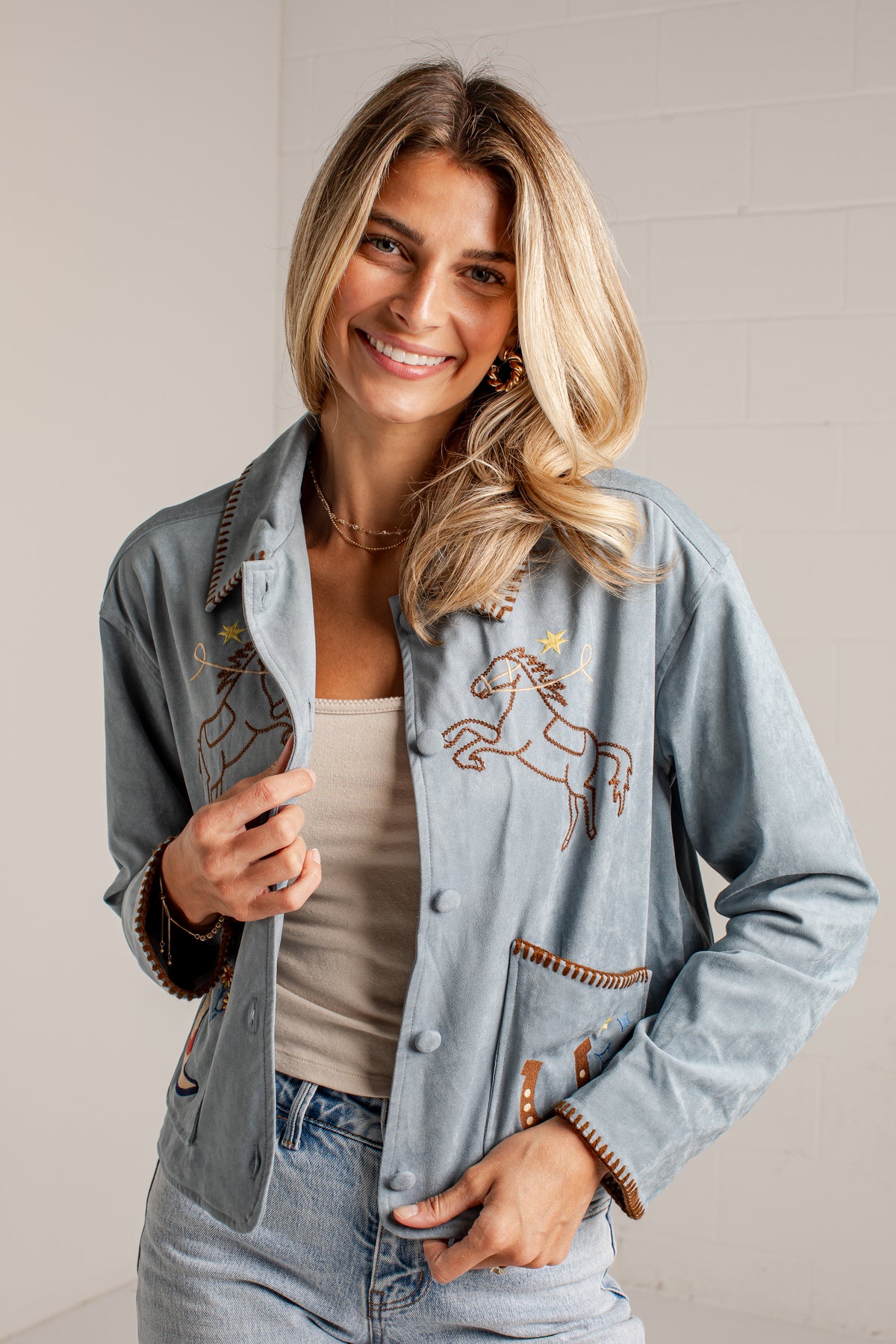 A close-up of a woman wearing a light blue suede jacket with western embroidery, including a horse design on the chest and a boot patch on the pocket.