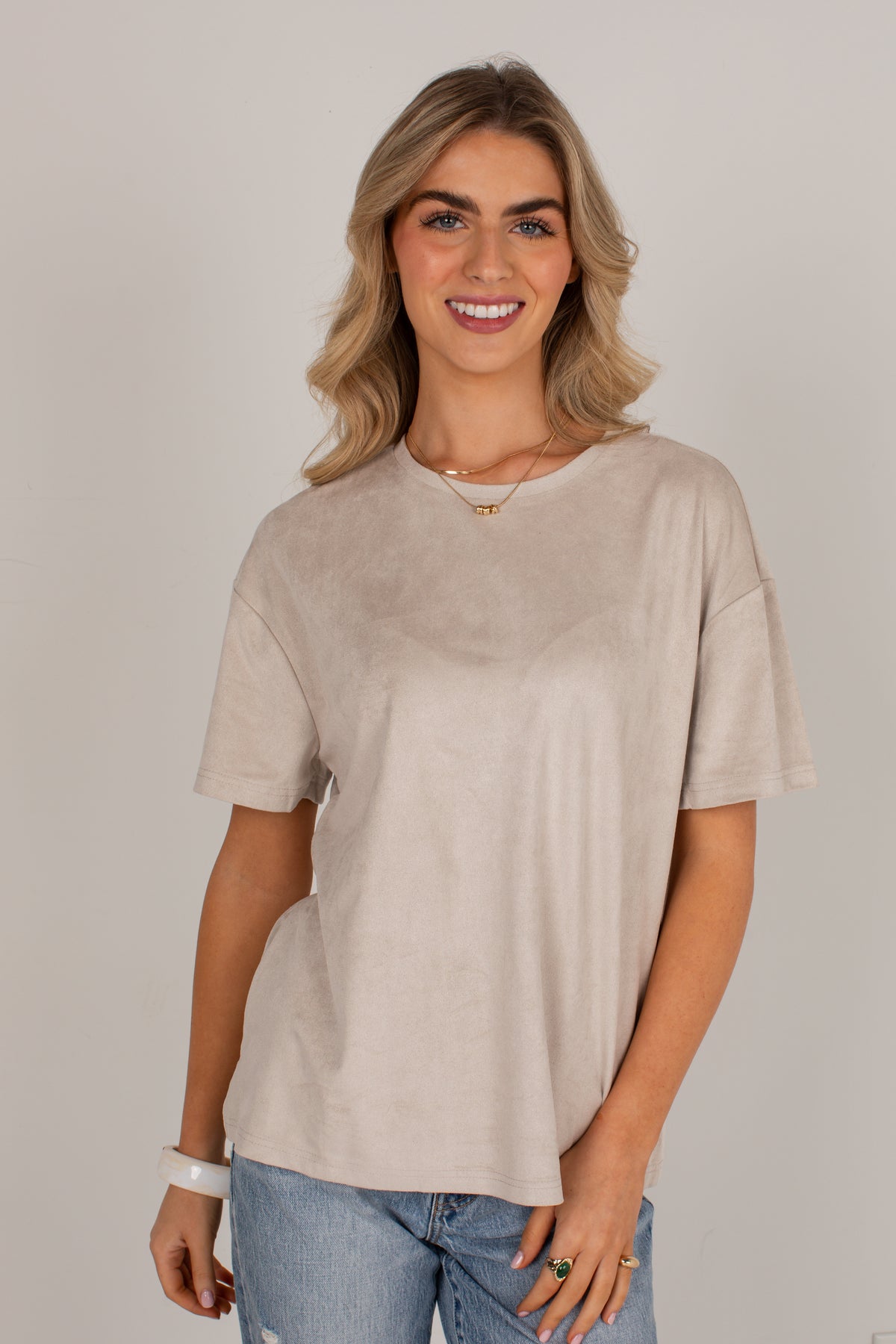 A woman with blonde hair wearing a light beige short-sleeve crewneck t-shirt and blue jeans, standing against a neutral light gray background.