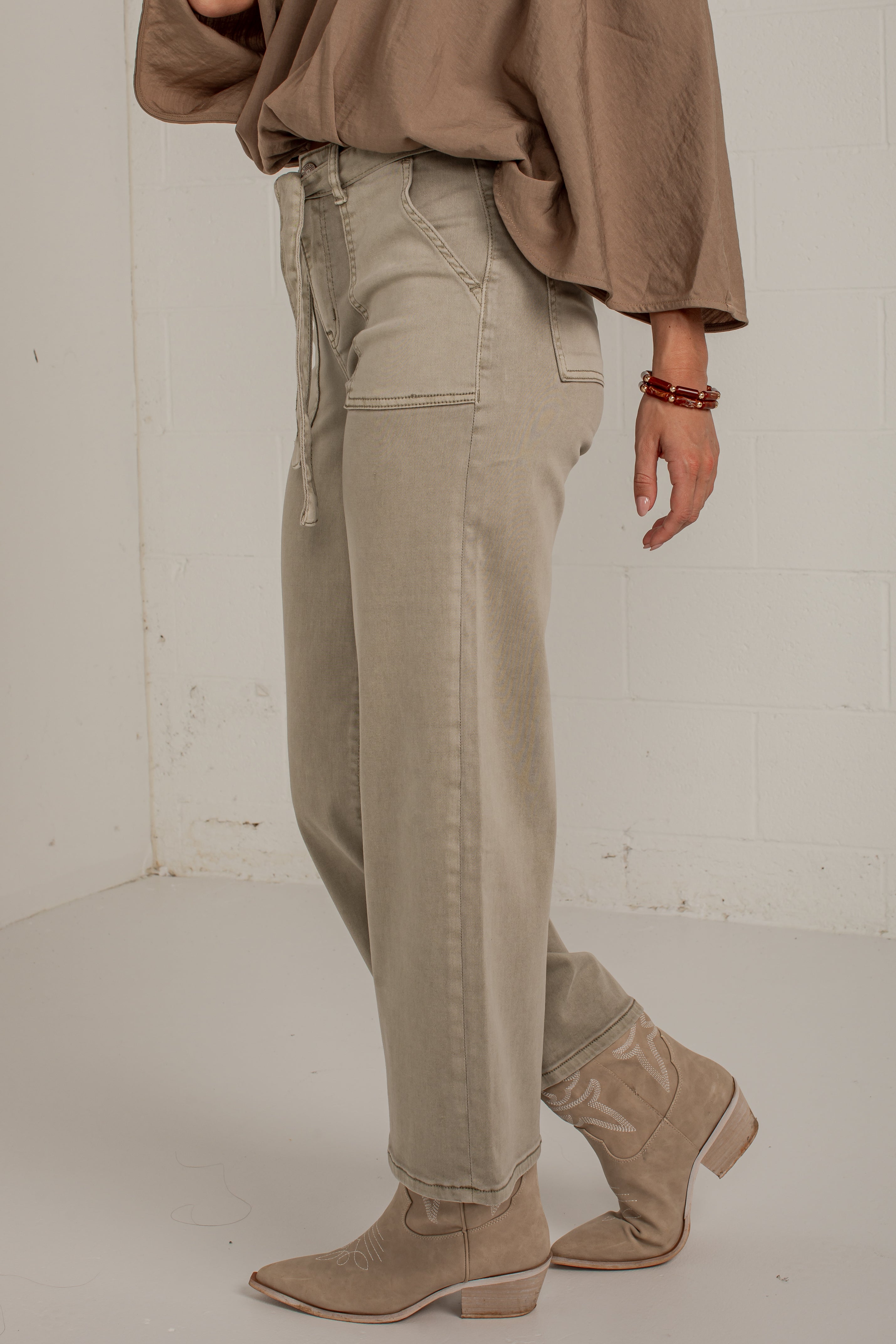 A person wearing light beige wide-leg jeans with a drawstring waist, paired with a taupe top and beige ankle boots, standing against a white brick wall.