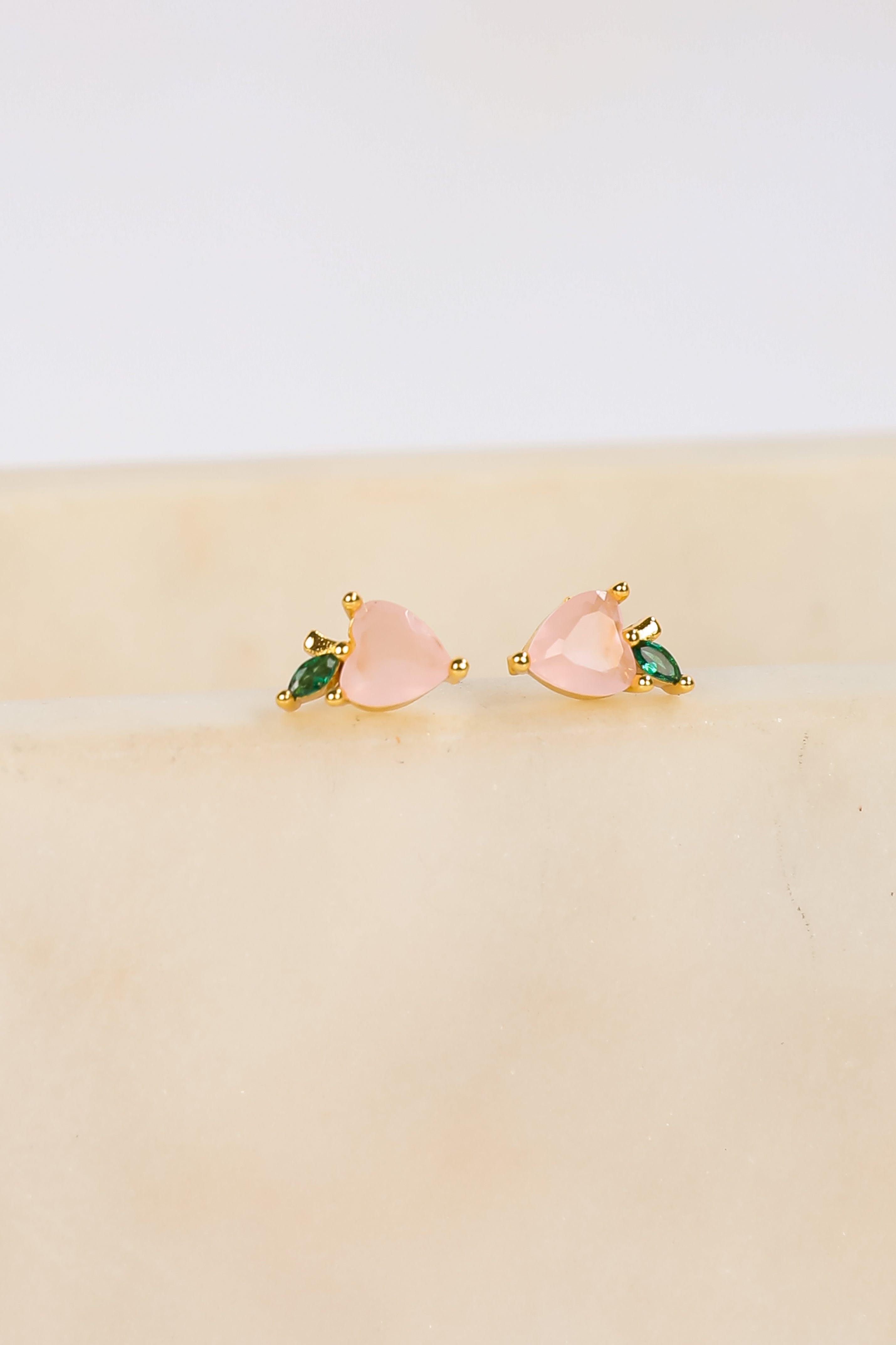 Pair of pink heart-shaped earrings with green gemstones on a beige surface.