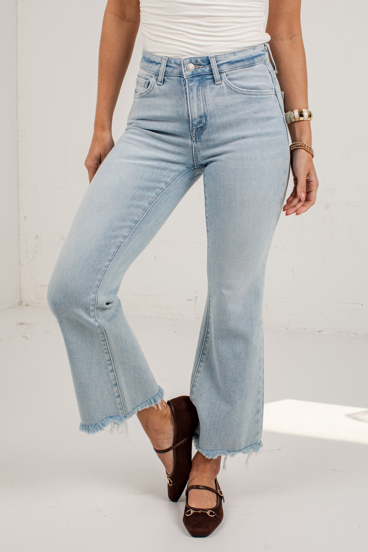 A model wearing light-wash blue flare jeans with a raw frayed hem, paired with a white top and dark brown flat shoes, standing against a white wall.
