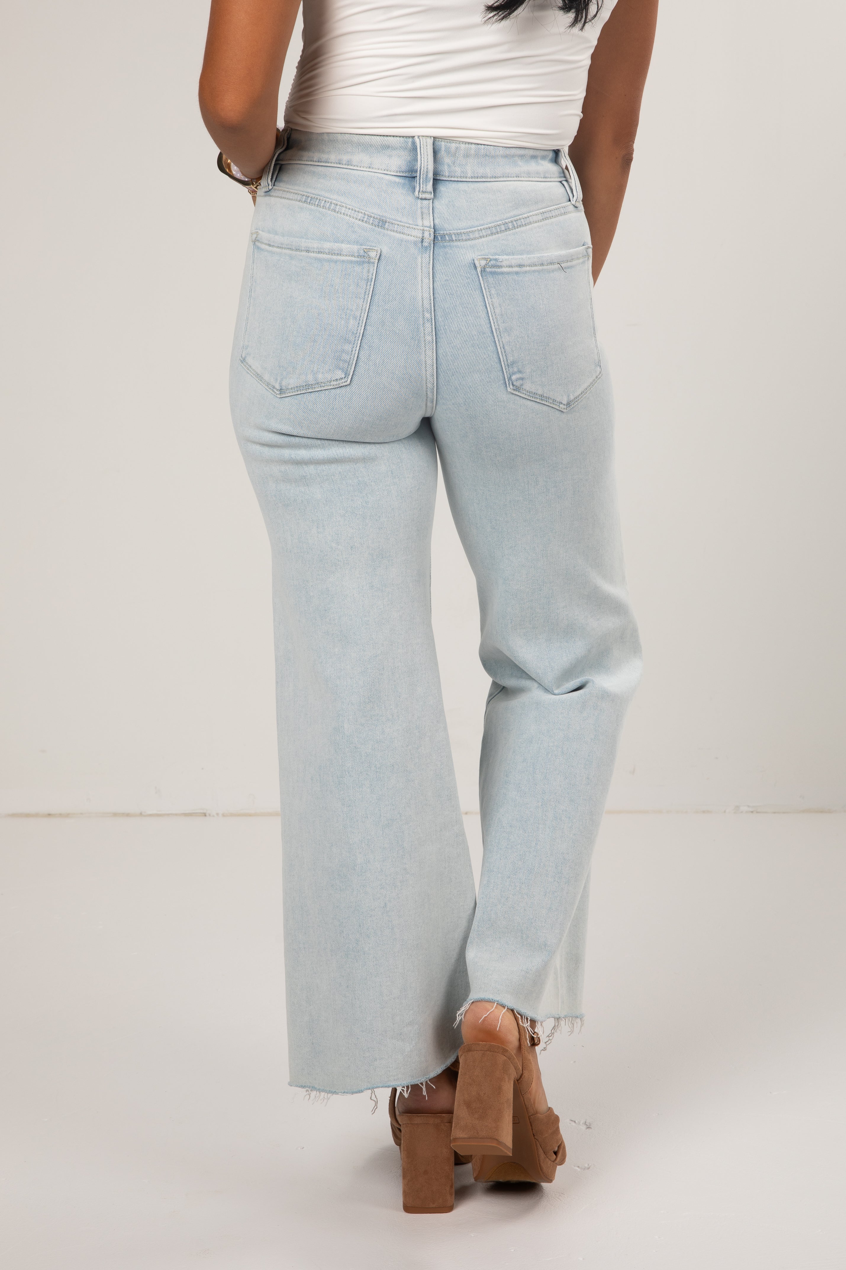 A person shown from the back wearing very light blue wide-leg jeans with a frayed hem, paired with a white top and brown block-heel sandals, against a plain off-white studio background.
