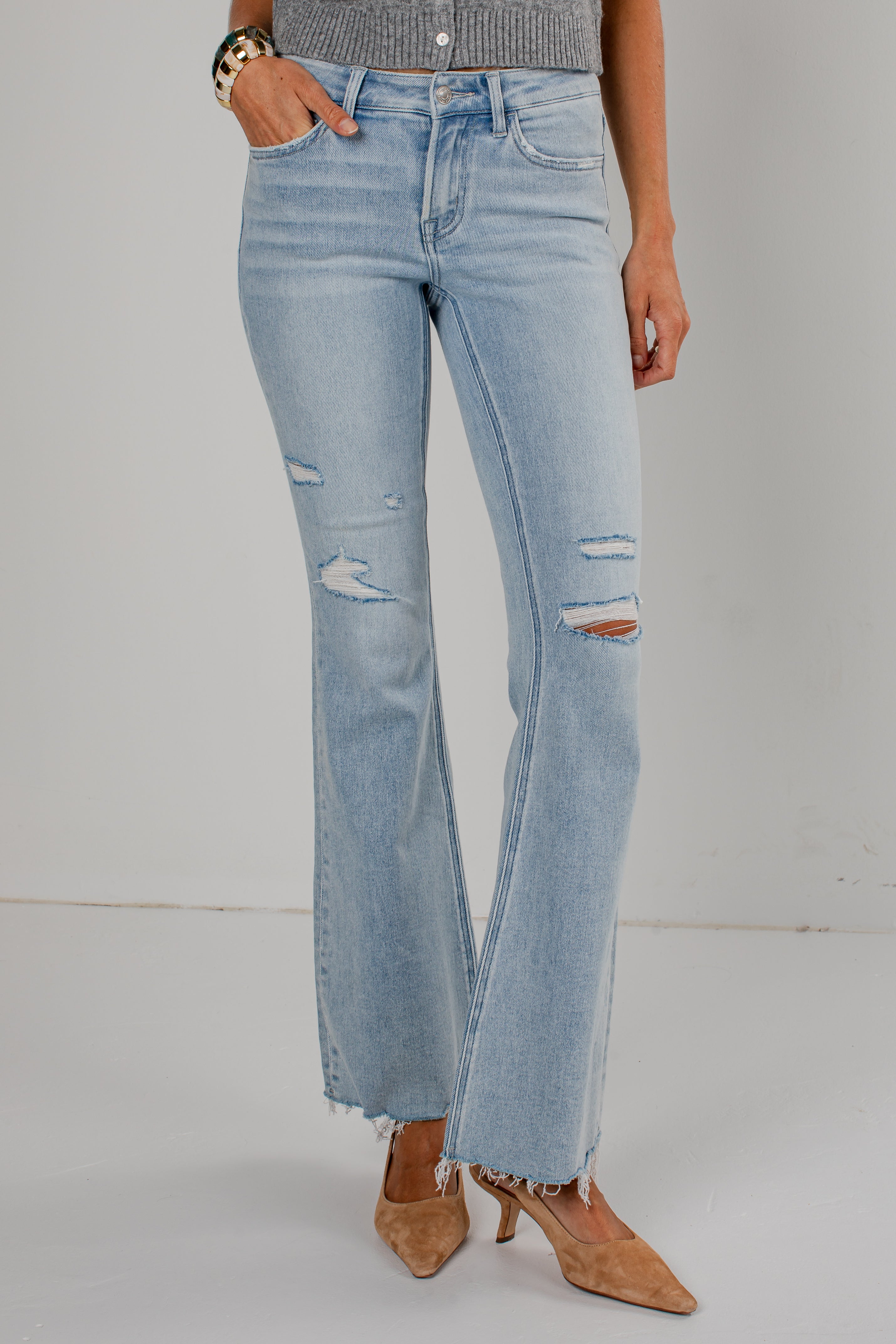 Light-wash blue distressed flare jeans with frayed hems and ripped details on the thighs and knees, shown on a model wearing tan pointed-toe heels against a light gray studio background.