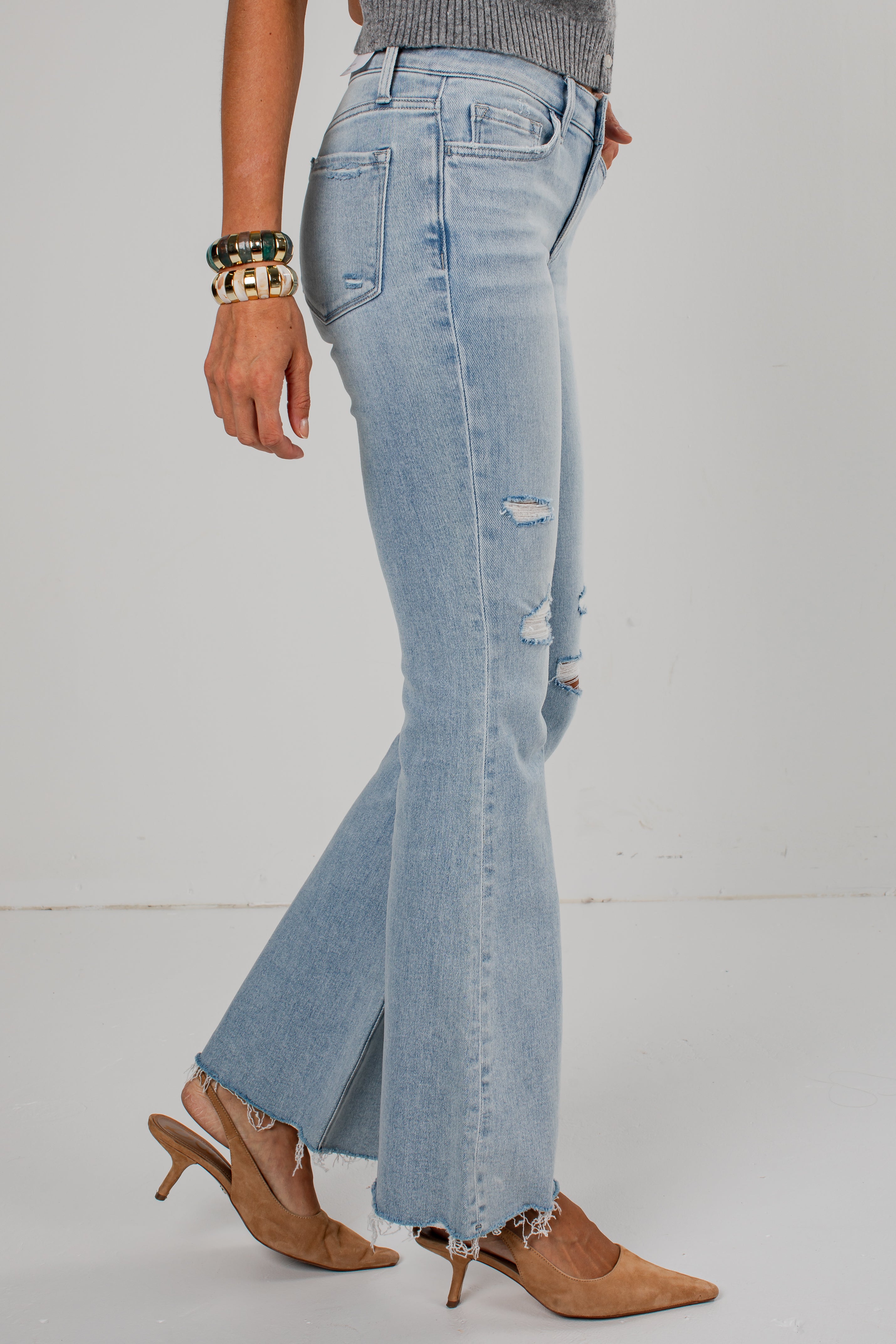 A person wearing light-wash blue distressed flare jeans with frayed hems, paired with tan pointed-toe heeled mules and bracelets, photographed against a plain light gray studio background.