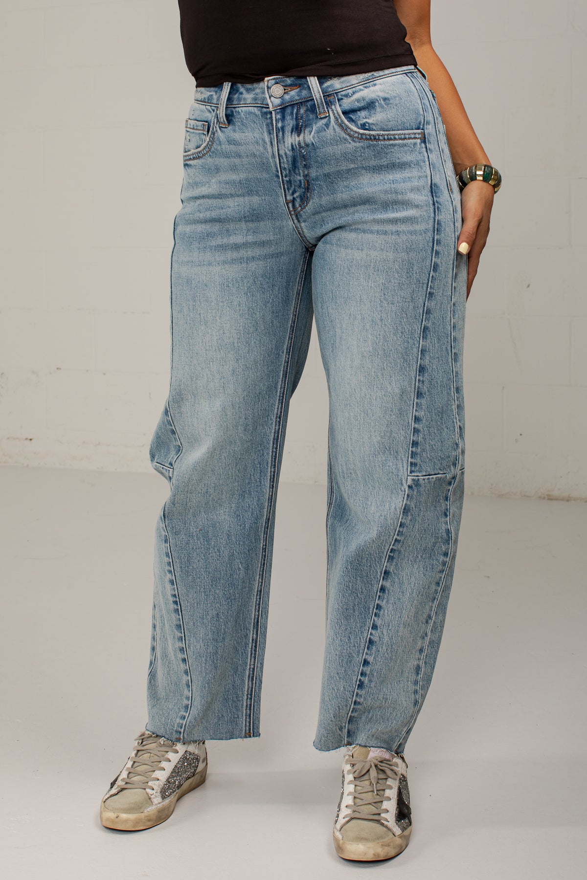 A person wearing light-wash blue wide-leg jeans with contrast stitching and side patch pockets, paired with a black top and beige sneakers, standing against a plain off-white wall.