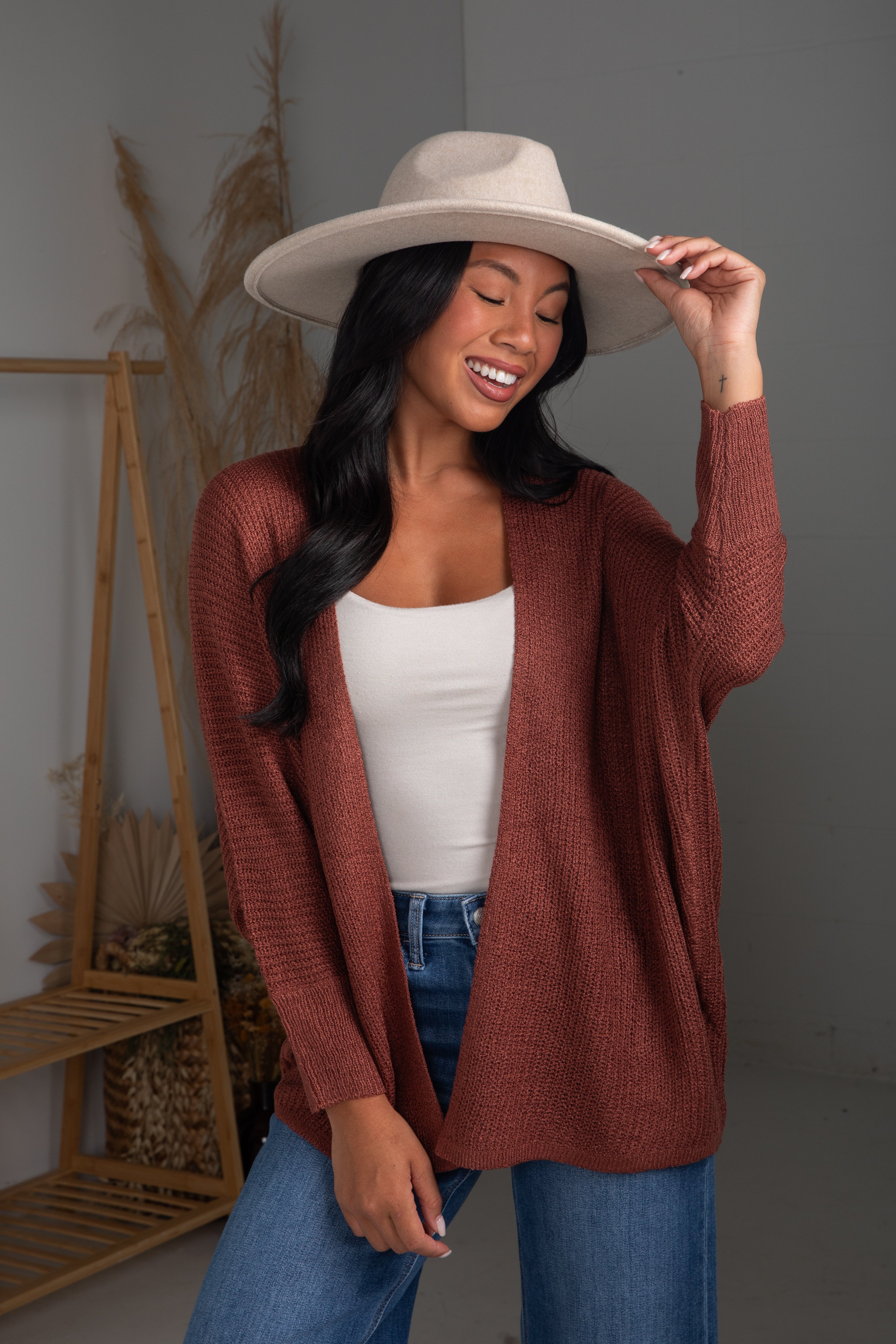 Woman wearing a rust-colored cardigan, white top, and blue jeans, holding a beige hat.