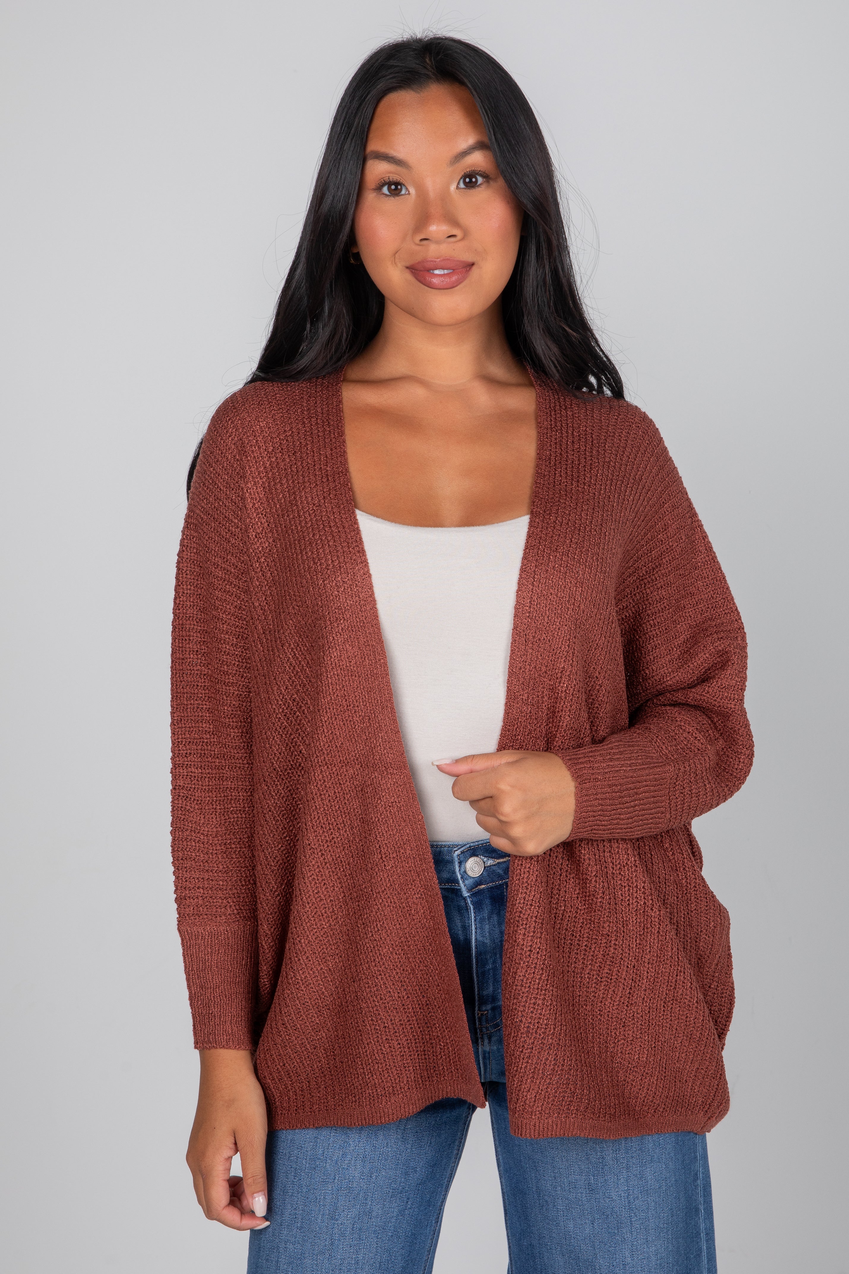 Woman wearing a brown cardigan over a white top and blue jeans on a gray background