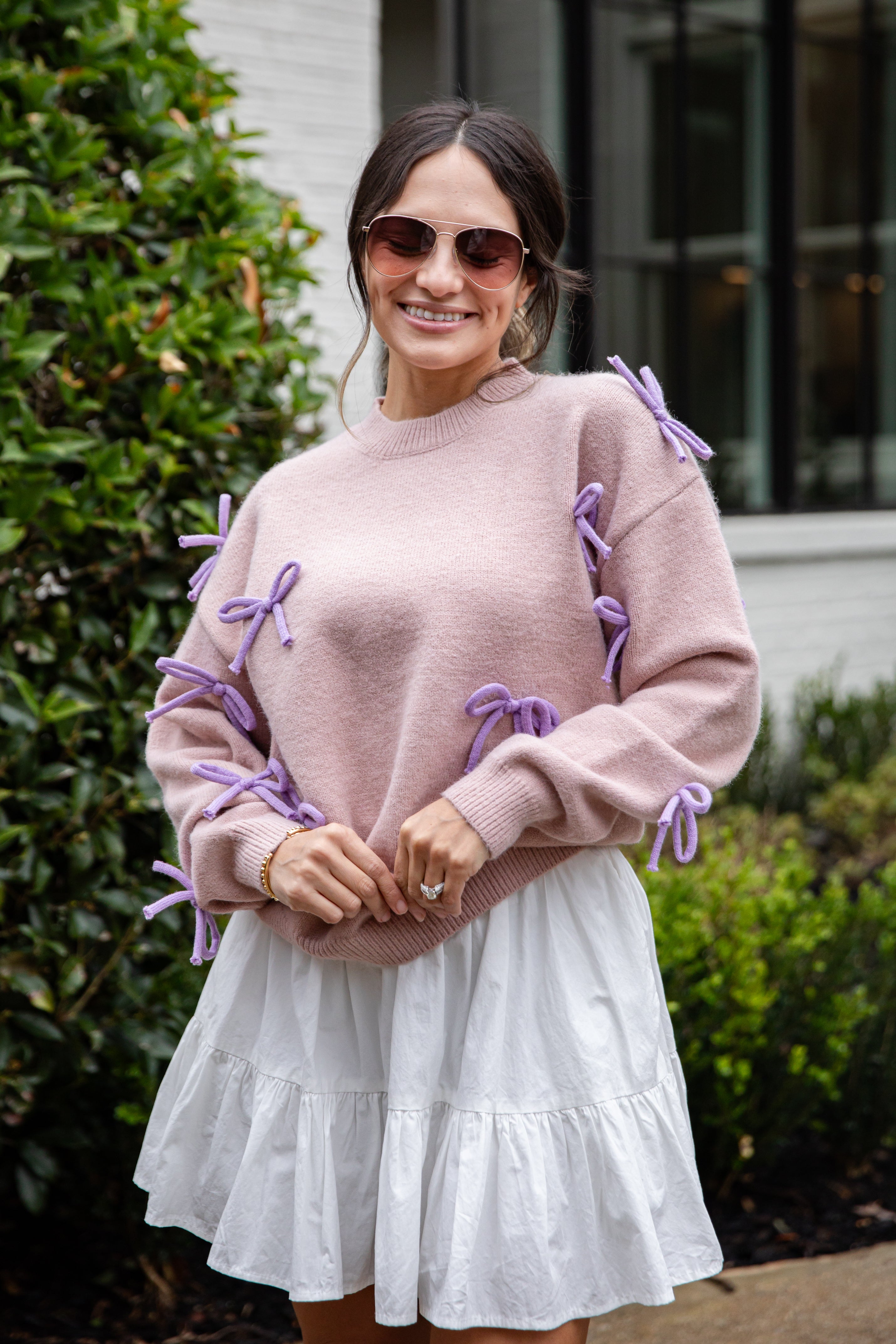 Woman wearing a pink sweater with purple accents and a white skirt outdoors.