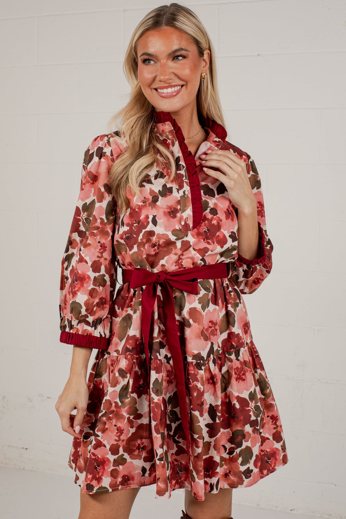 A smiling blonde woman models a short floral dress with a red collar, cuffs, and waist tie, set against a white brick wall.