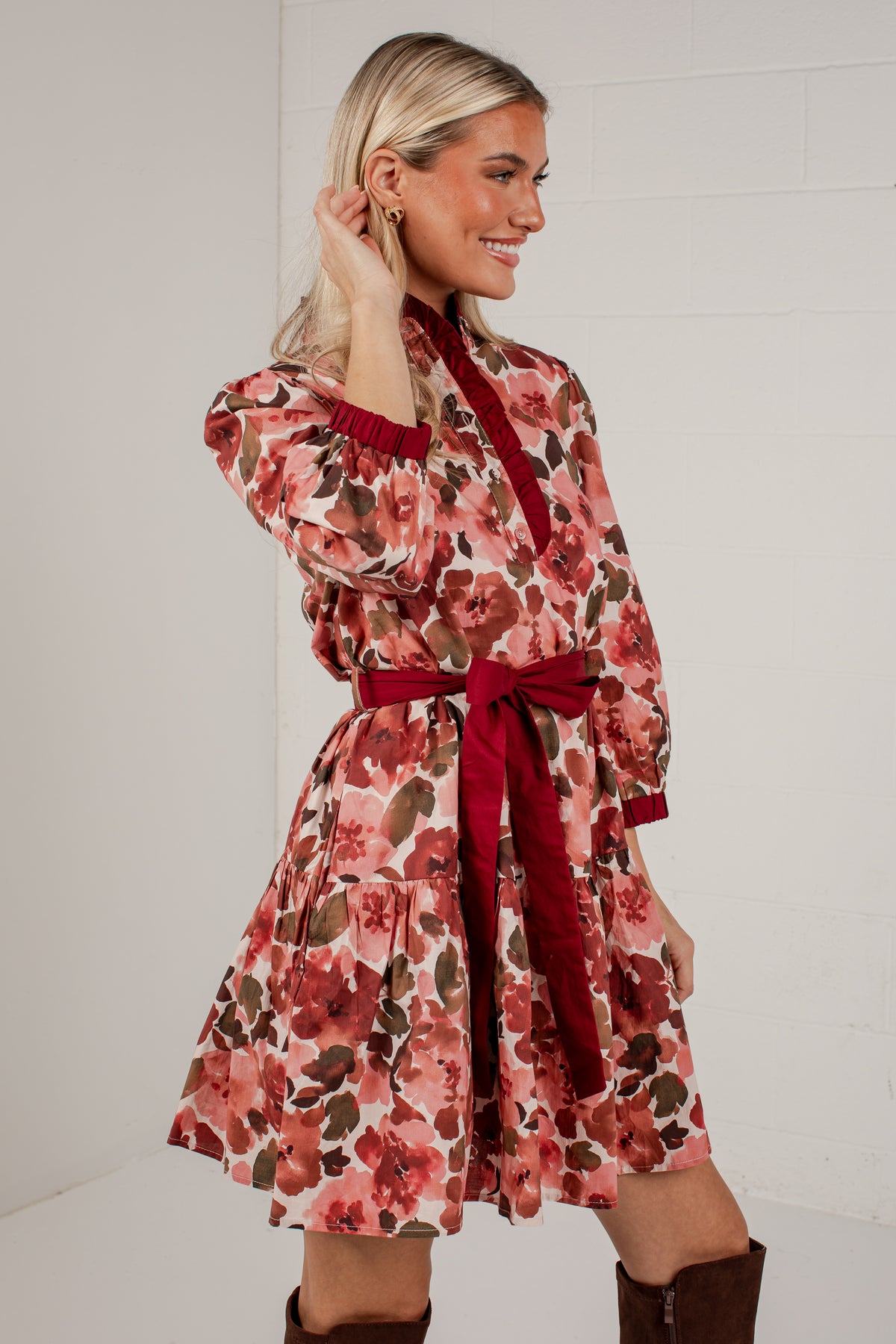 A woman modeling a long-sleeve floral mini dress with a dark red tie belt, shown against a white brick wall background.