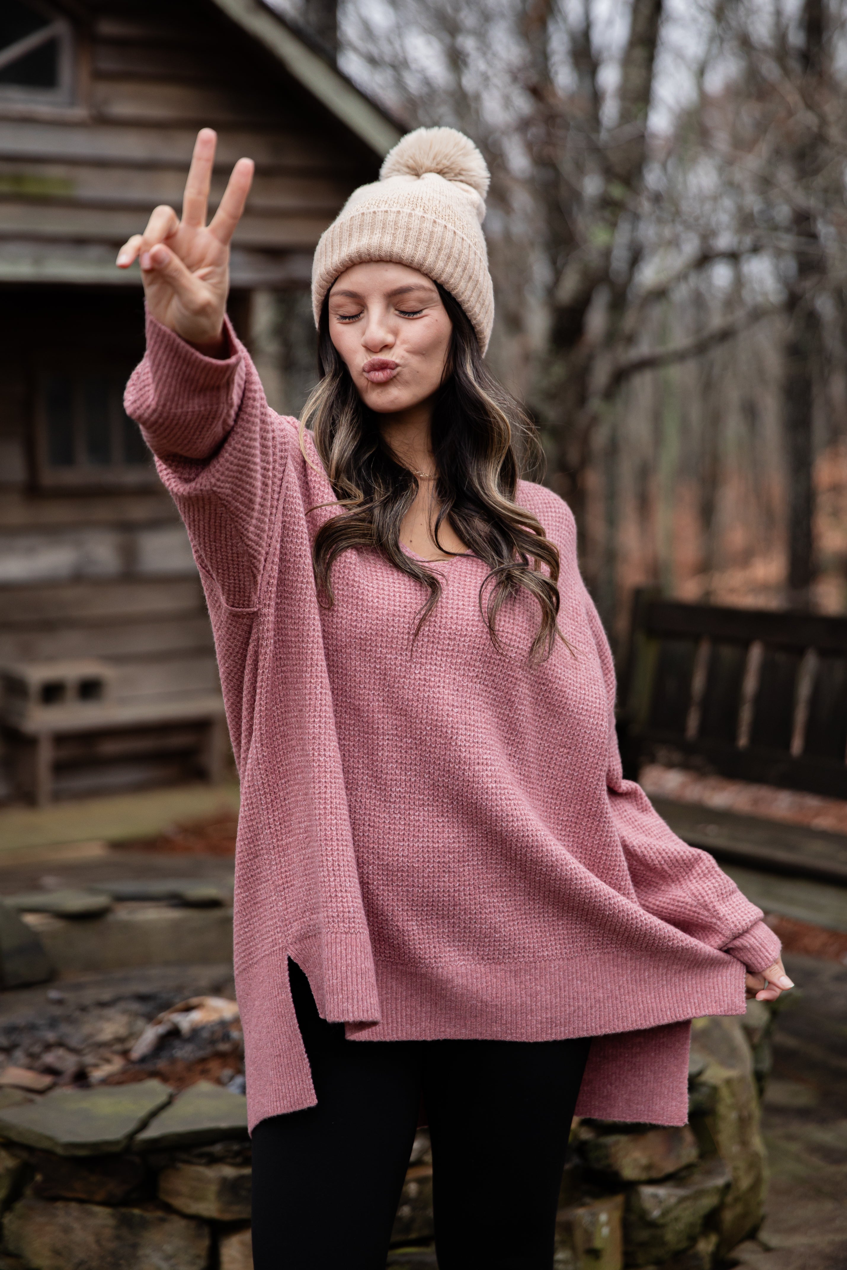 Woman in a pink sweater and beige beanie making a peace sign outdoors.