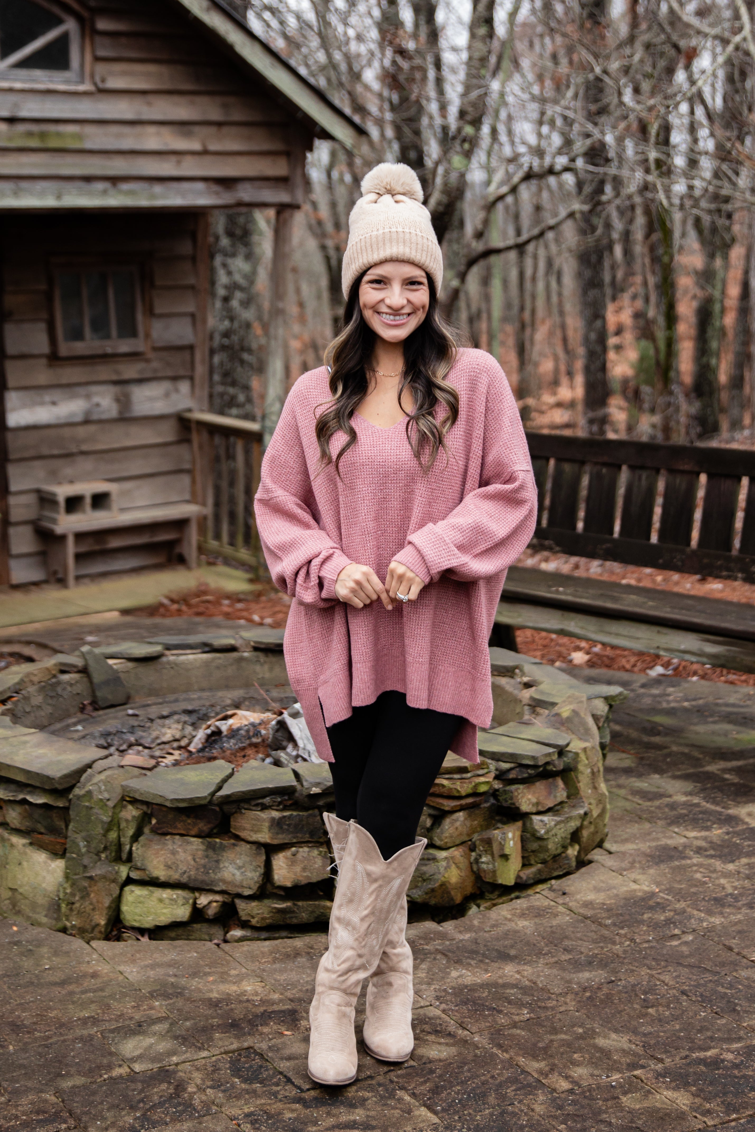 Woman in pink sweater and white boots standing in front of a wooden cabin.