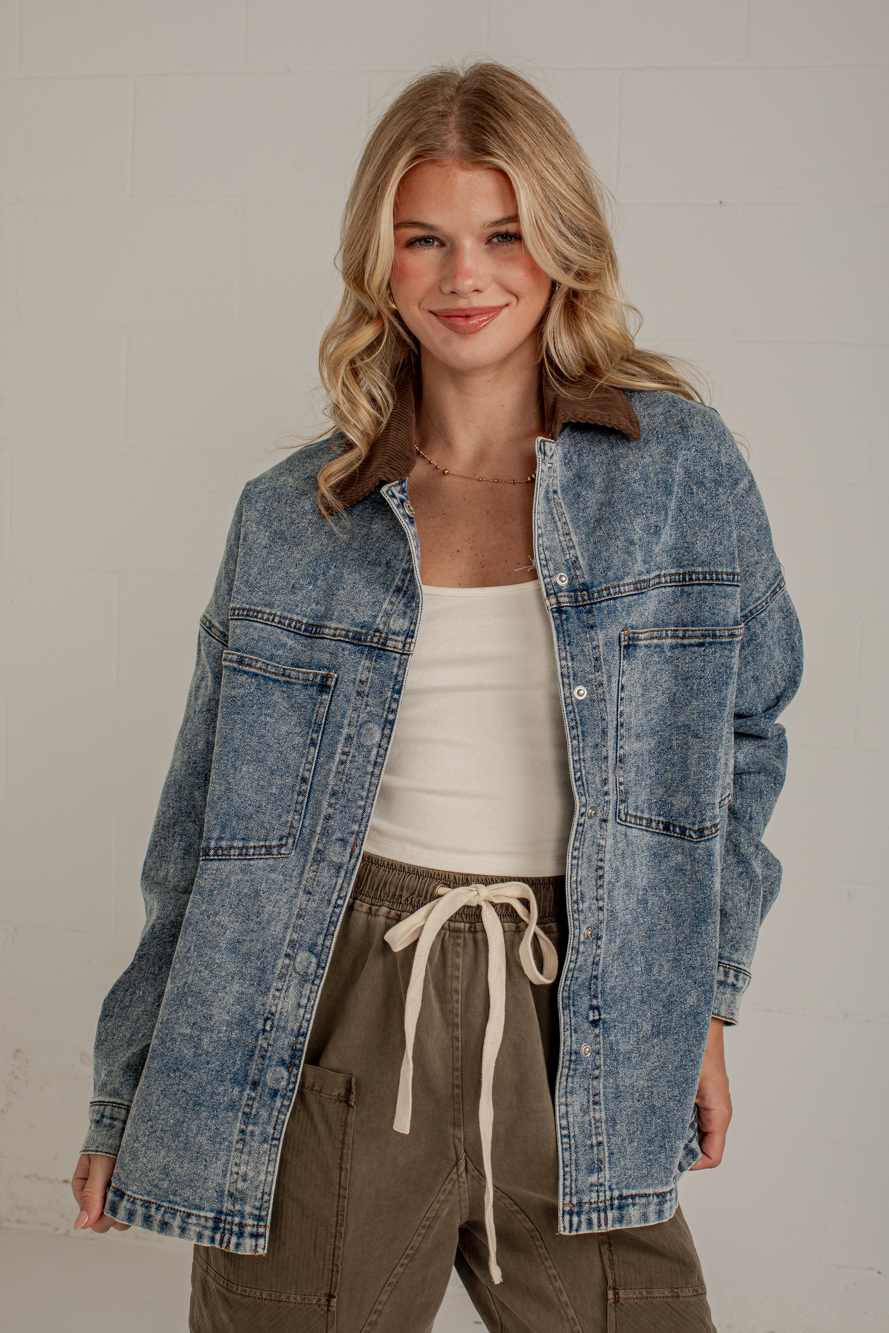 A woman wearing a light blue acid-wash denim shirt jacket with a brown collar, styled over a white tank top and olive green drawstring cargo pants, standing against a light gray brick wall.