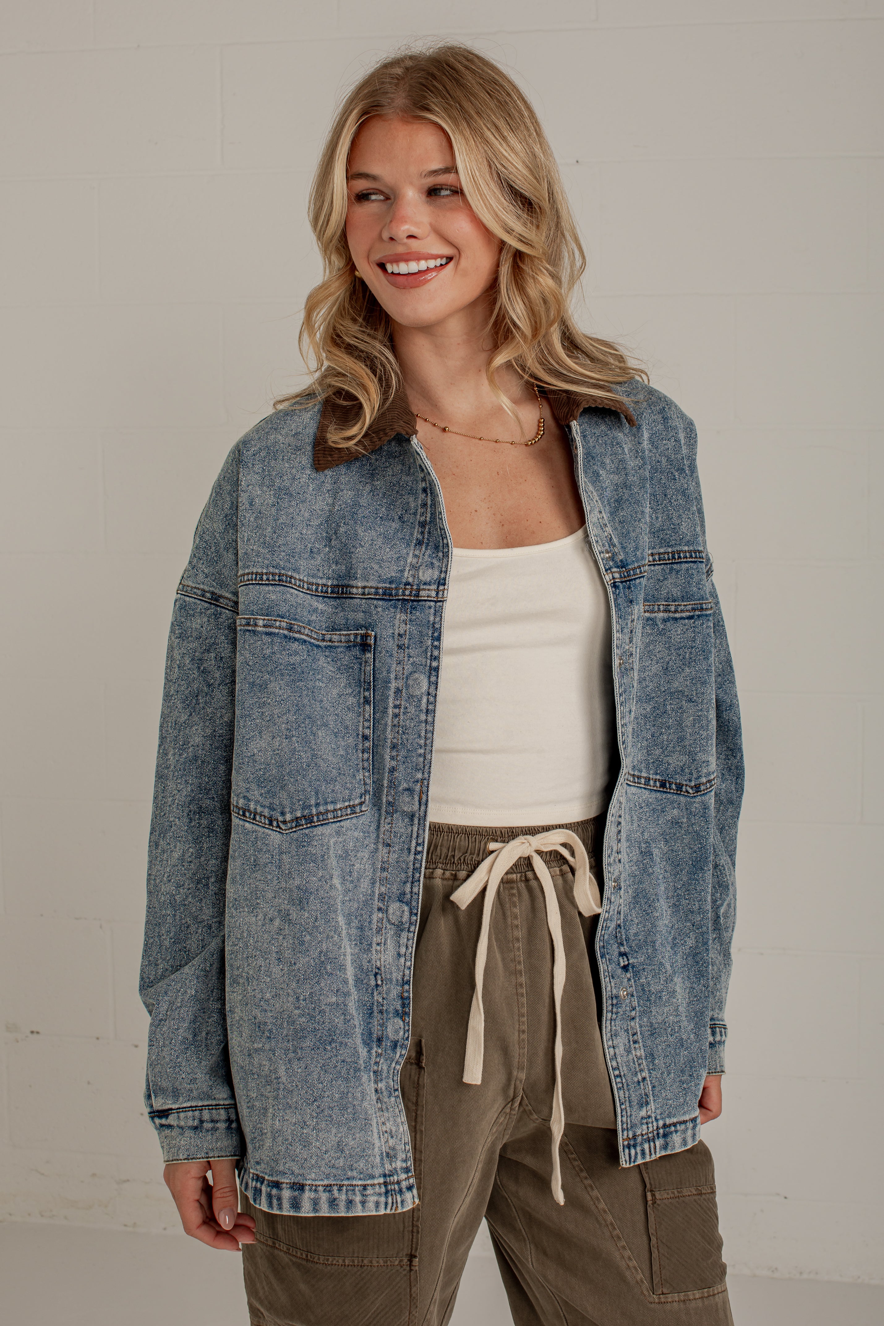 A woman wearing a light blue acid-wash denim jacket over a white tank top and olive green drawstring cargo pants, standing against a light gray wall.