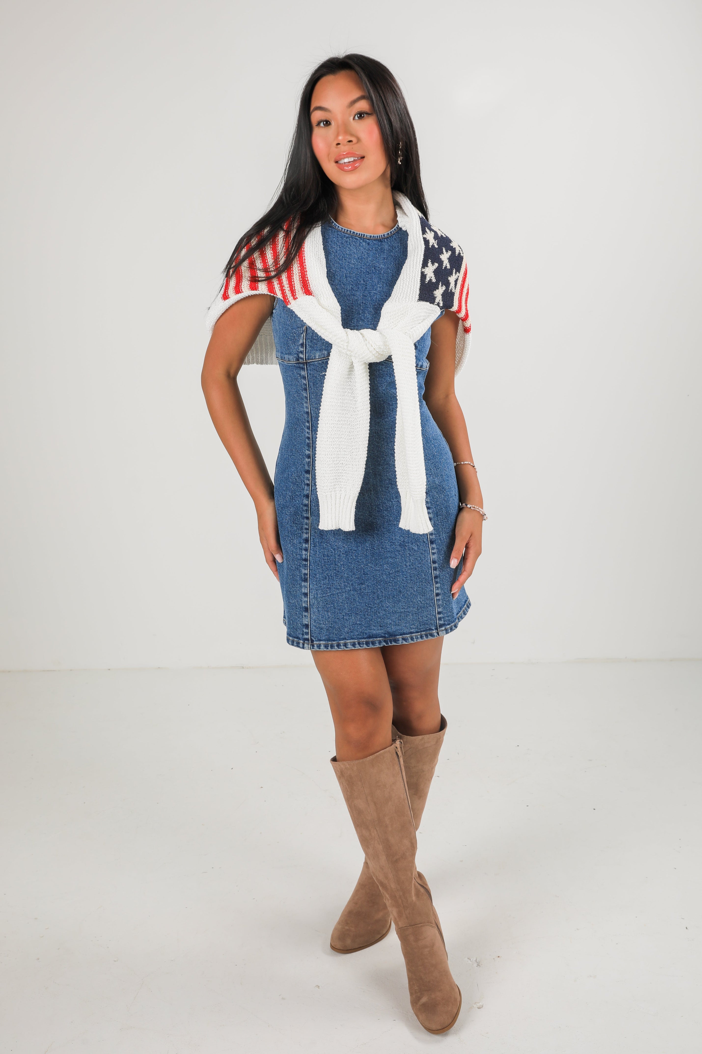 Woman wearing a denim dress with an American flag scarf and brown boots on a white background