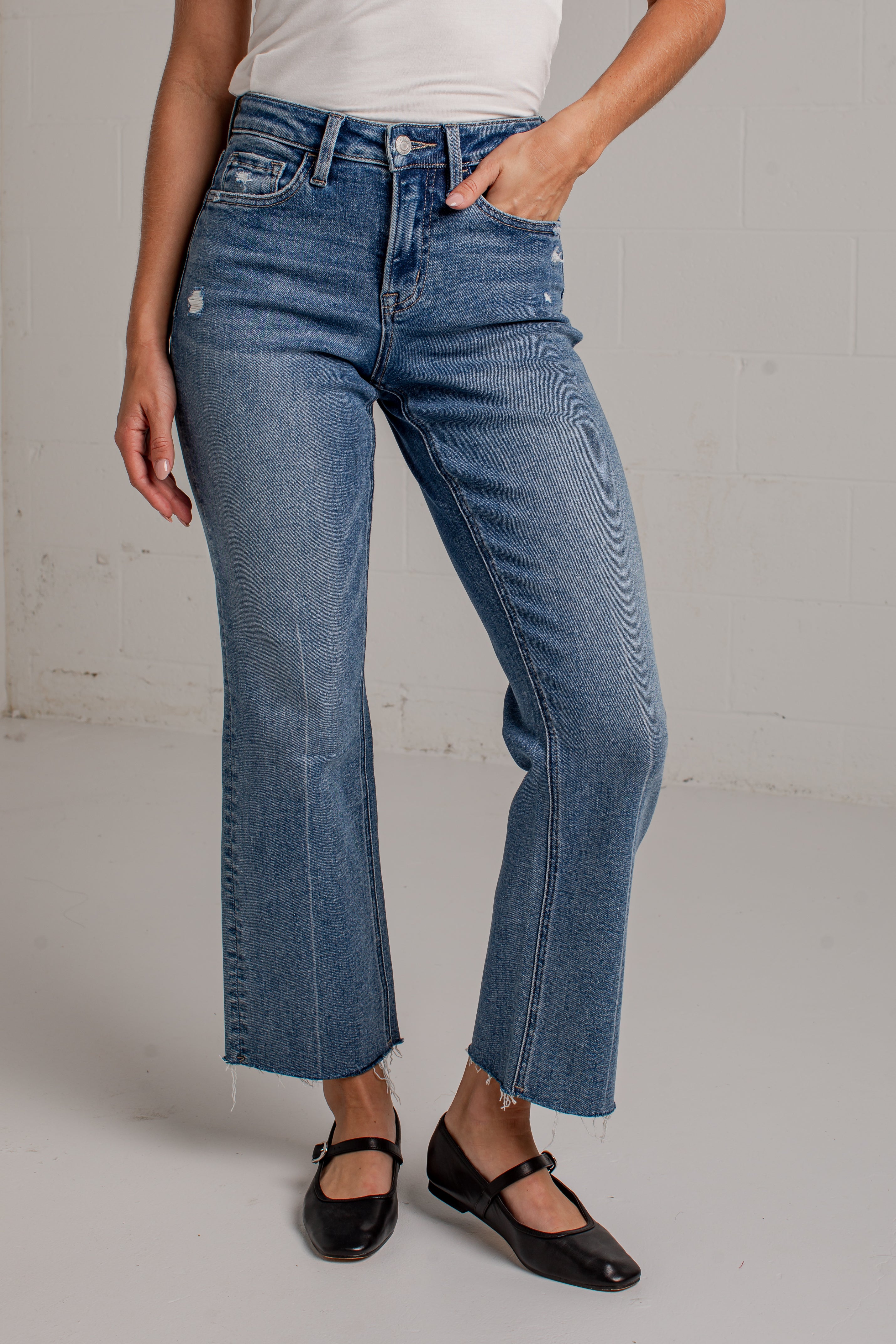A woman modeling a white short-sleeve scoop-neck top and blue cropped wide leg jeans with black flat shoes, standing against a white brick wall.