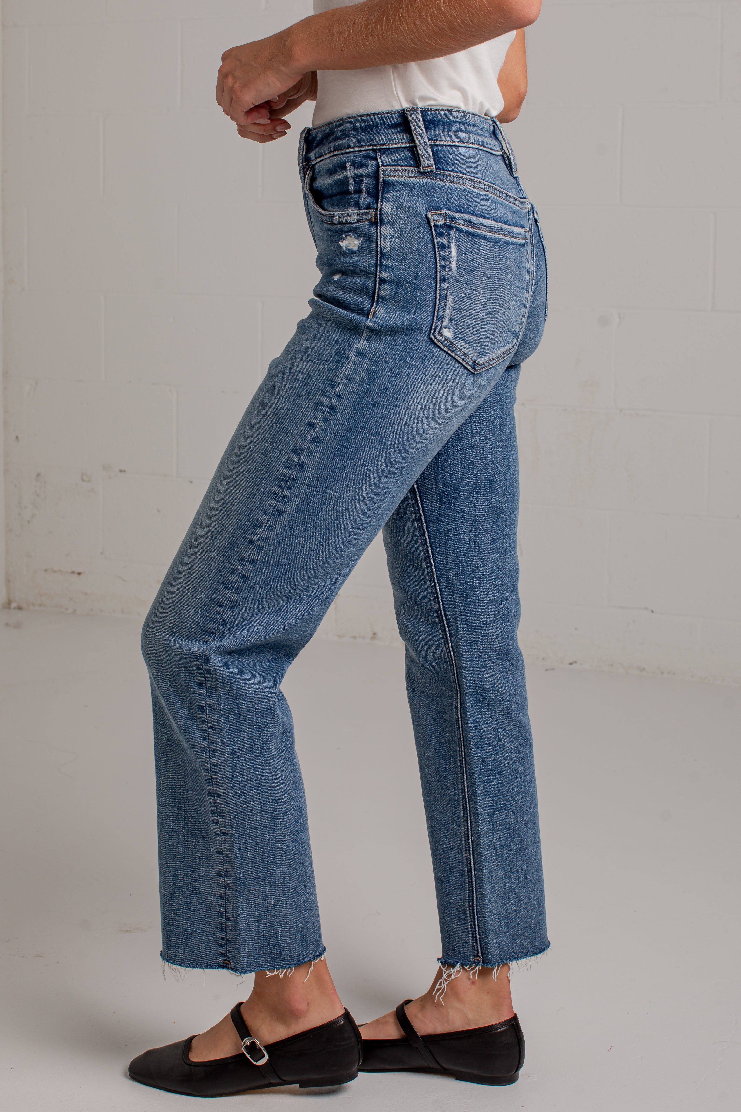 A person wearing medium-wash blue denim jeans with a straight-to-crew leg, frayed hems, and distressed details on the back pockets, paired with black flat shoes against a white brick wall background.