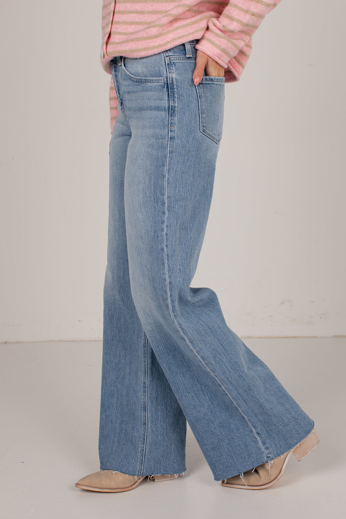 A person wearing light-wash blue wide-leg flare jeans, a pink and white striped long-sleeve top, and beige pointed-toe flats, standing against a plain off-white studio background.