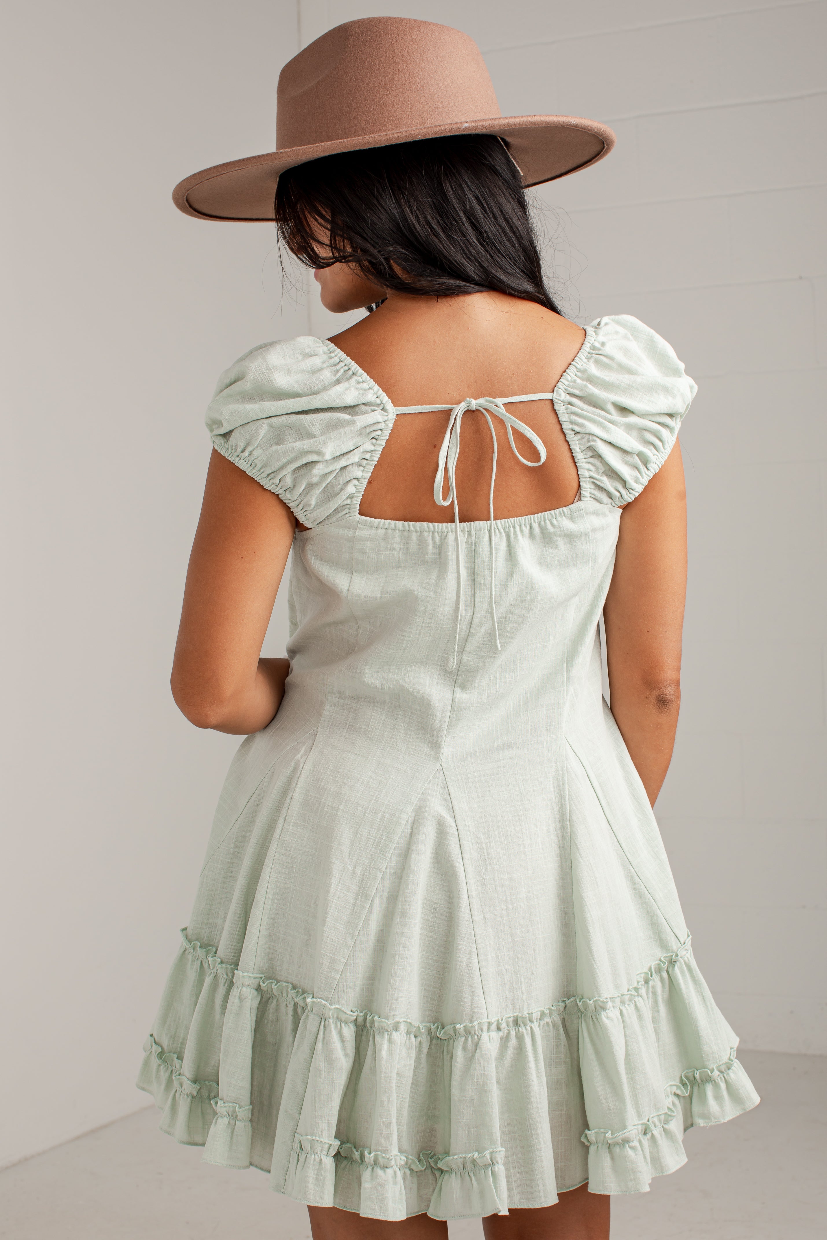 A woman shown from the back wearing a pale mint green puff-sleeve dress with a tie-back neckline and a matching wide-brim tan hat, set against a light gray wall.