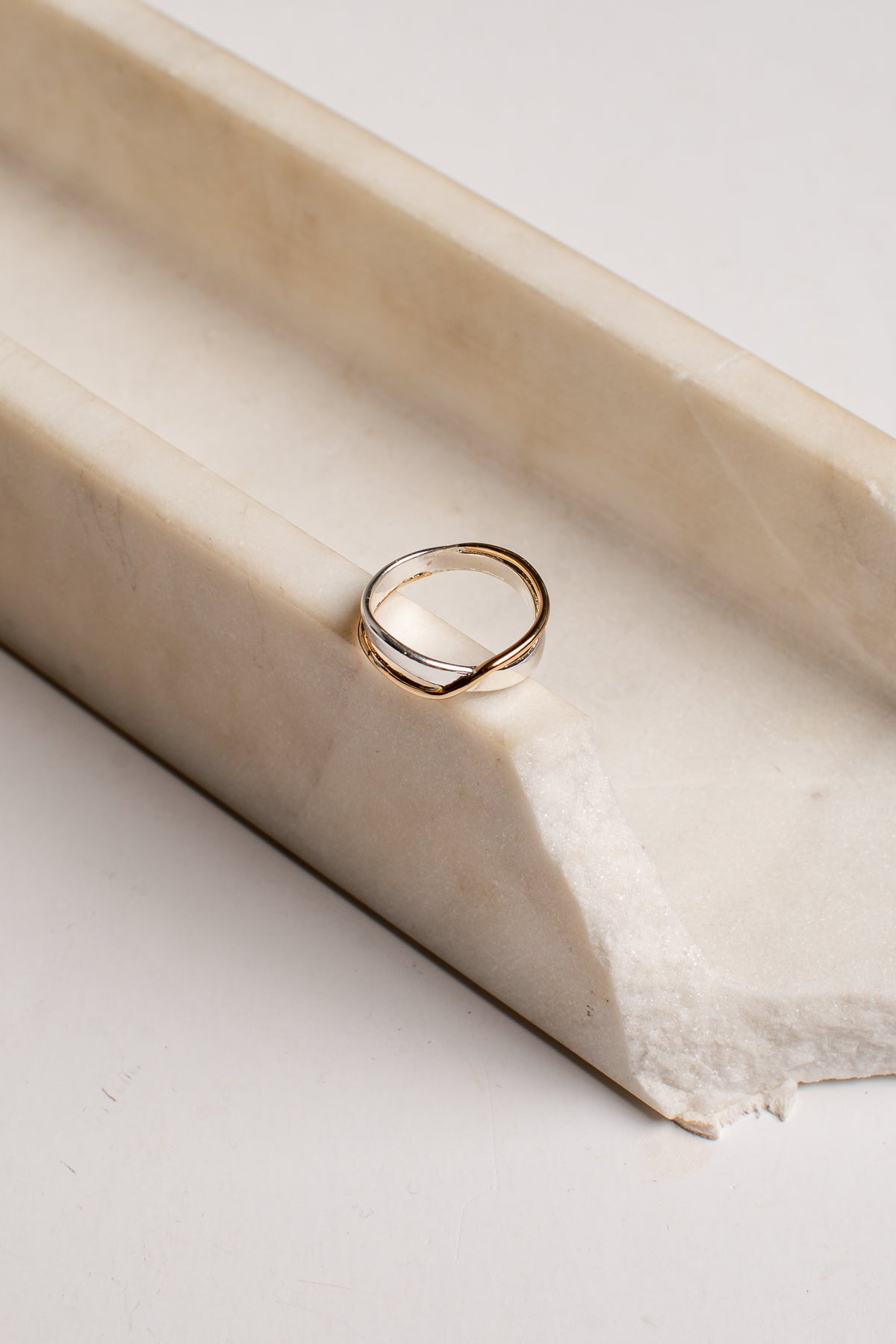 A thin double-band ring with a twisted, intertwined design in polished yellow gold, resting on a cream-colored stone riser against a light background.