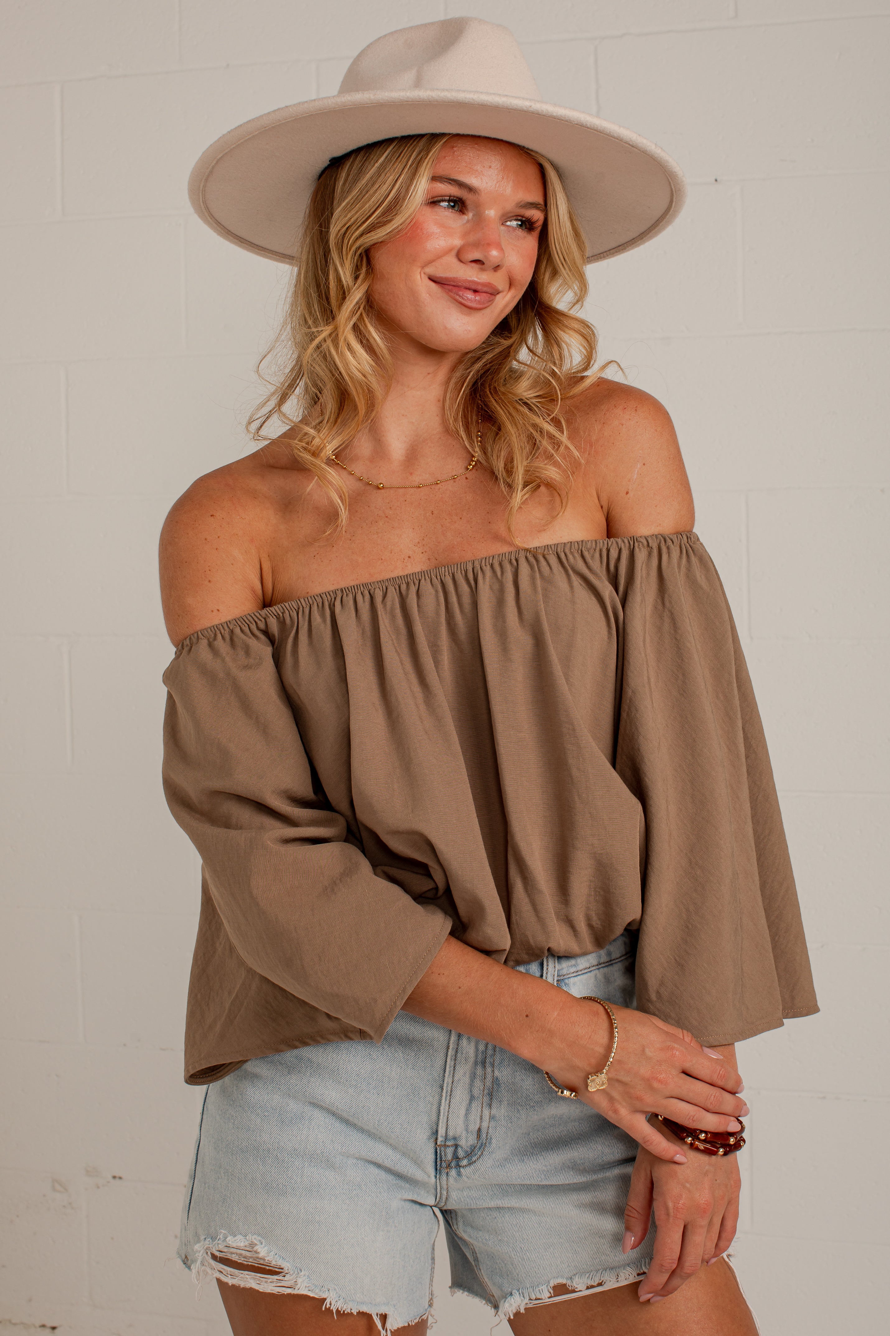 A woman wearing a taupe brown off-the-shoulder top with three-quarter sleeves, paired with light-wash denim shorts and a beige wide-brim hat, posing against a white brick wall.