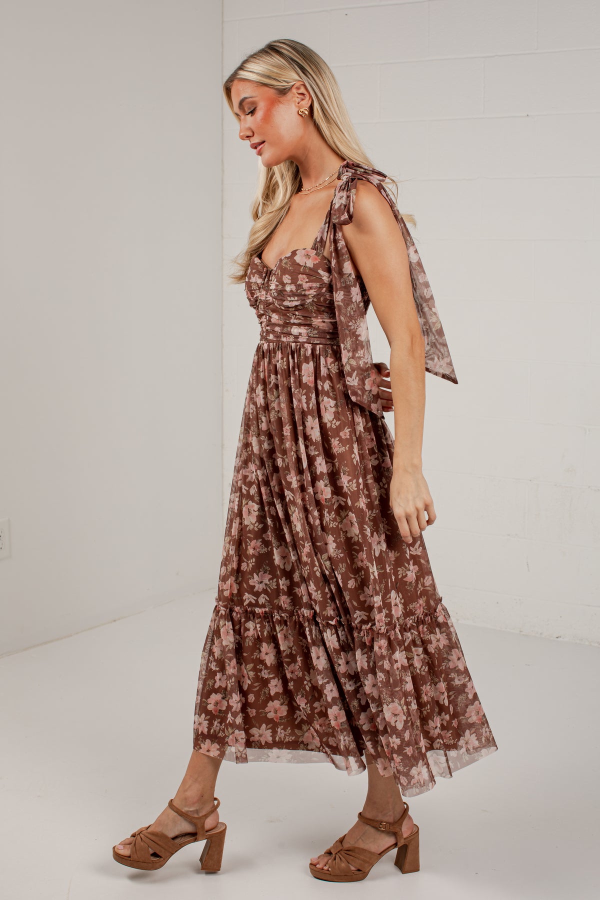 A woman modeling a brown floral midi dress with tie-shoulder straps and a matching waist tie, paired with brown heeled sandals, set against a white brick wall background.