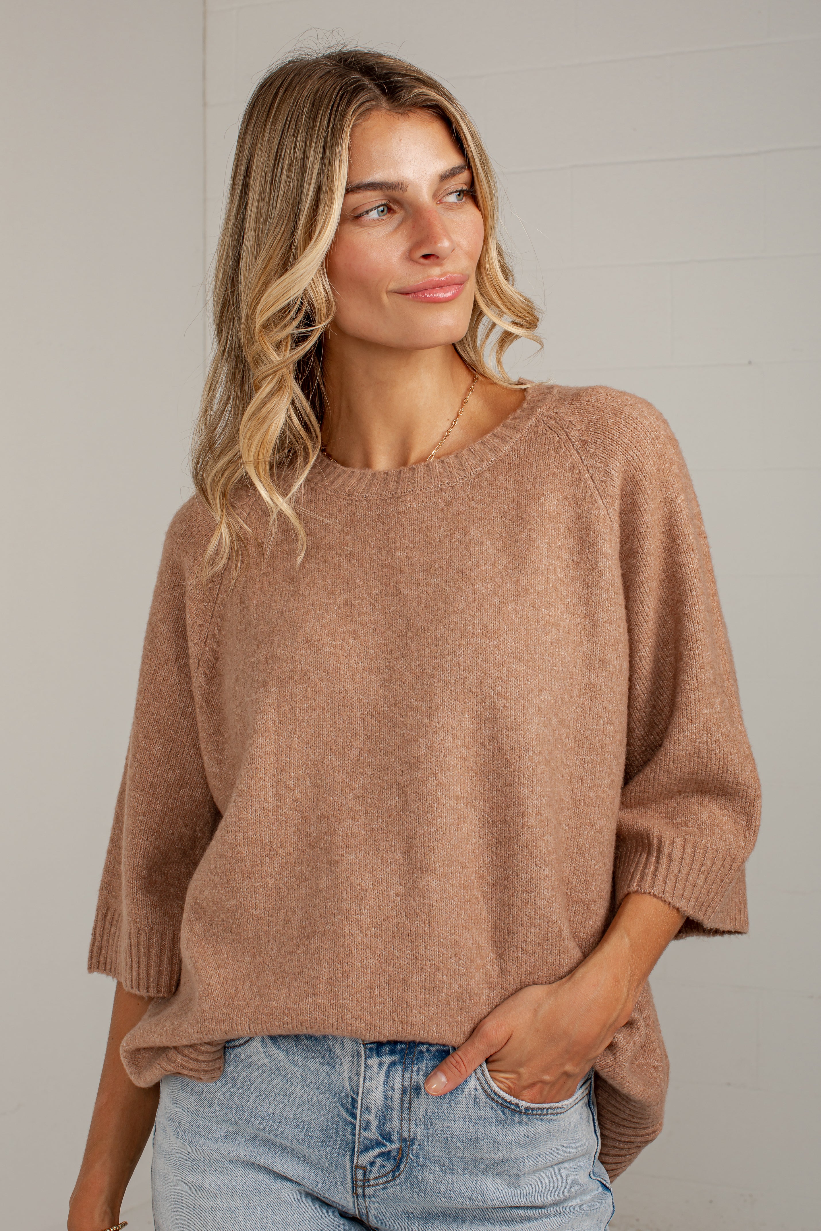 A woman wearing a soft, heathered taupe-brown knit sweater with elbow-length sleeves and a relaxed fit, paired with light-wash blue jeans, standing against a light gray wall.