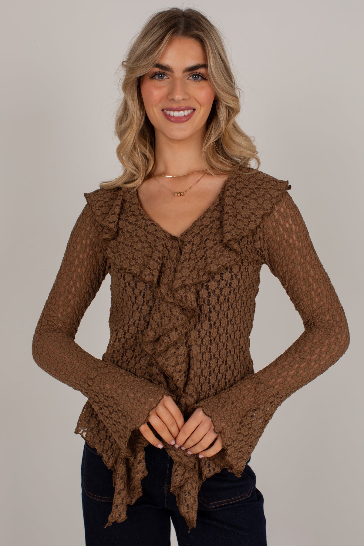 A smiling blonde woman wearing a brown textured knit long-sleeve top with ruffle details and dark blue jeans, standing against a plain light gray studio background.