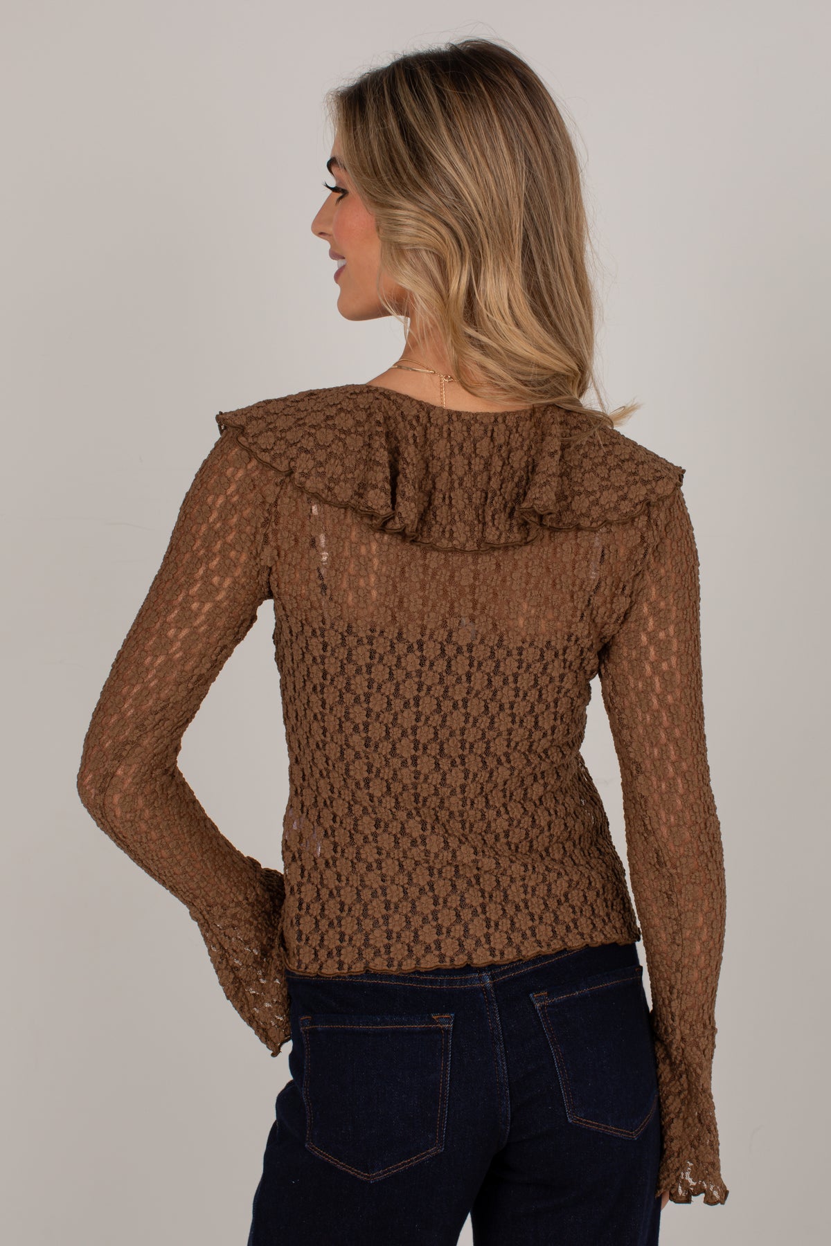 A woman shown from the back wearing a brown textured long-sleeve top with a ruffled collar and dark blue jeans, against a plain light gray studio background.
