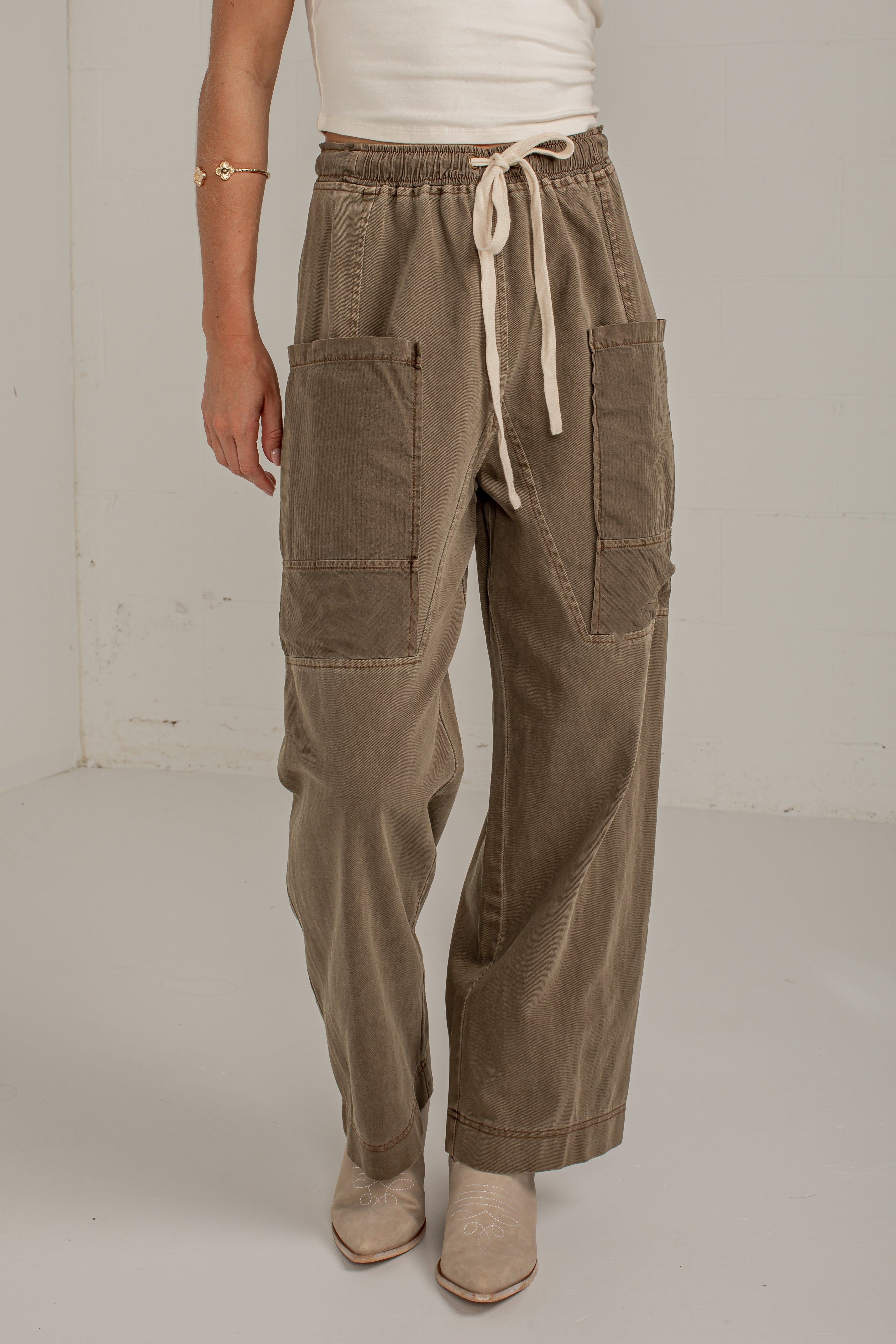 A person wearing wide-leg taupe corduroy pants with a cream drawstring waist, large patch pockets, and beige ankle boots, standing against a plain off-white studio wall.