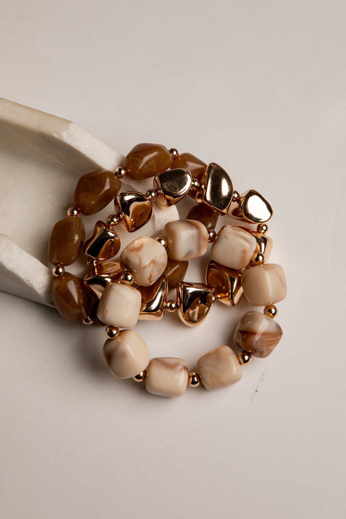 A set of three stretch bracelets with chunky, organic-shaped beads in shades of brown, cream, and marbled beige, separated by small metallic gold spacer beads. One bracelet is displayed on a white ceramic stand against a light gray background.