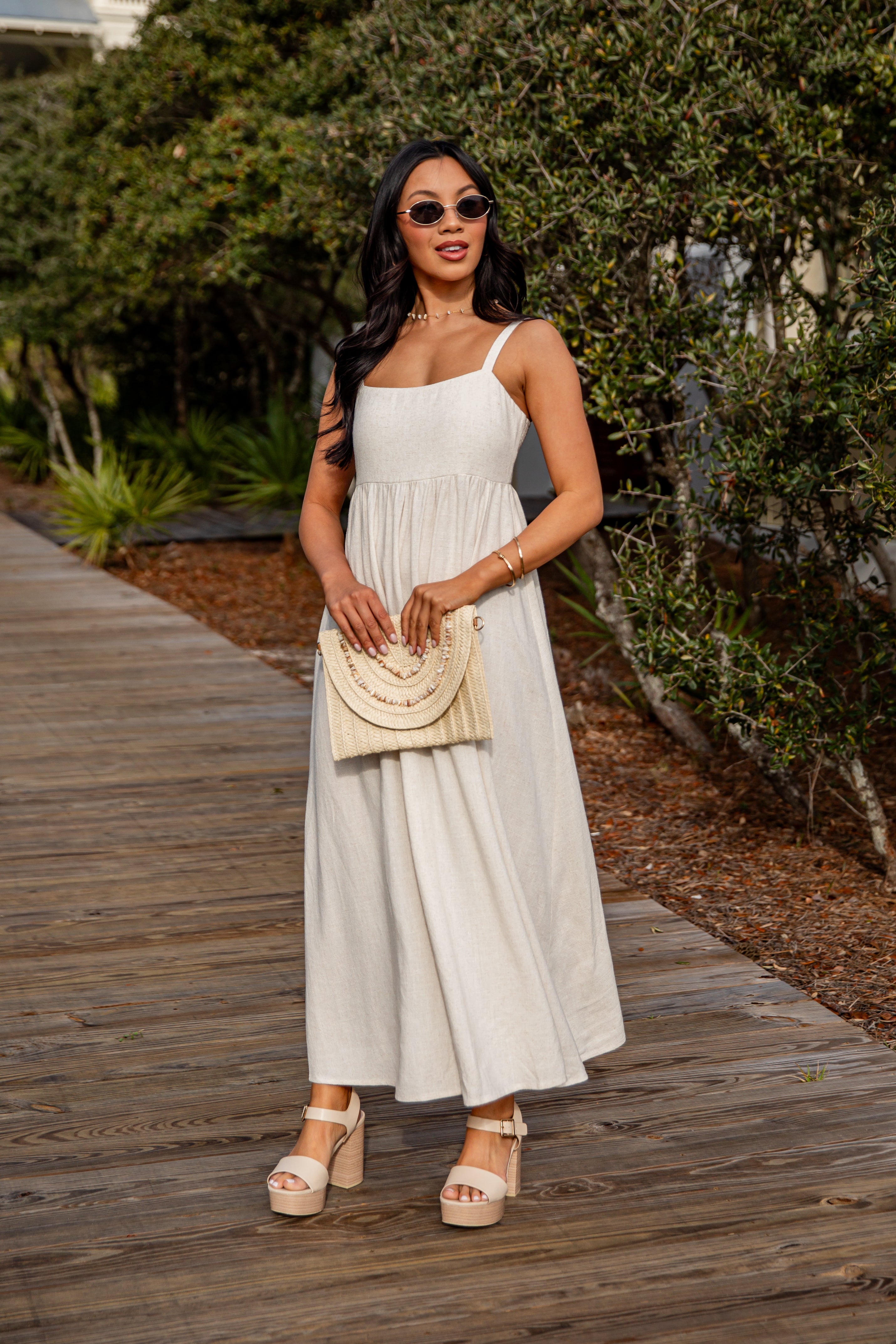 Woman in a white dress holding a beige handbag on a wooden path with greenery.