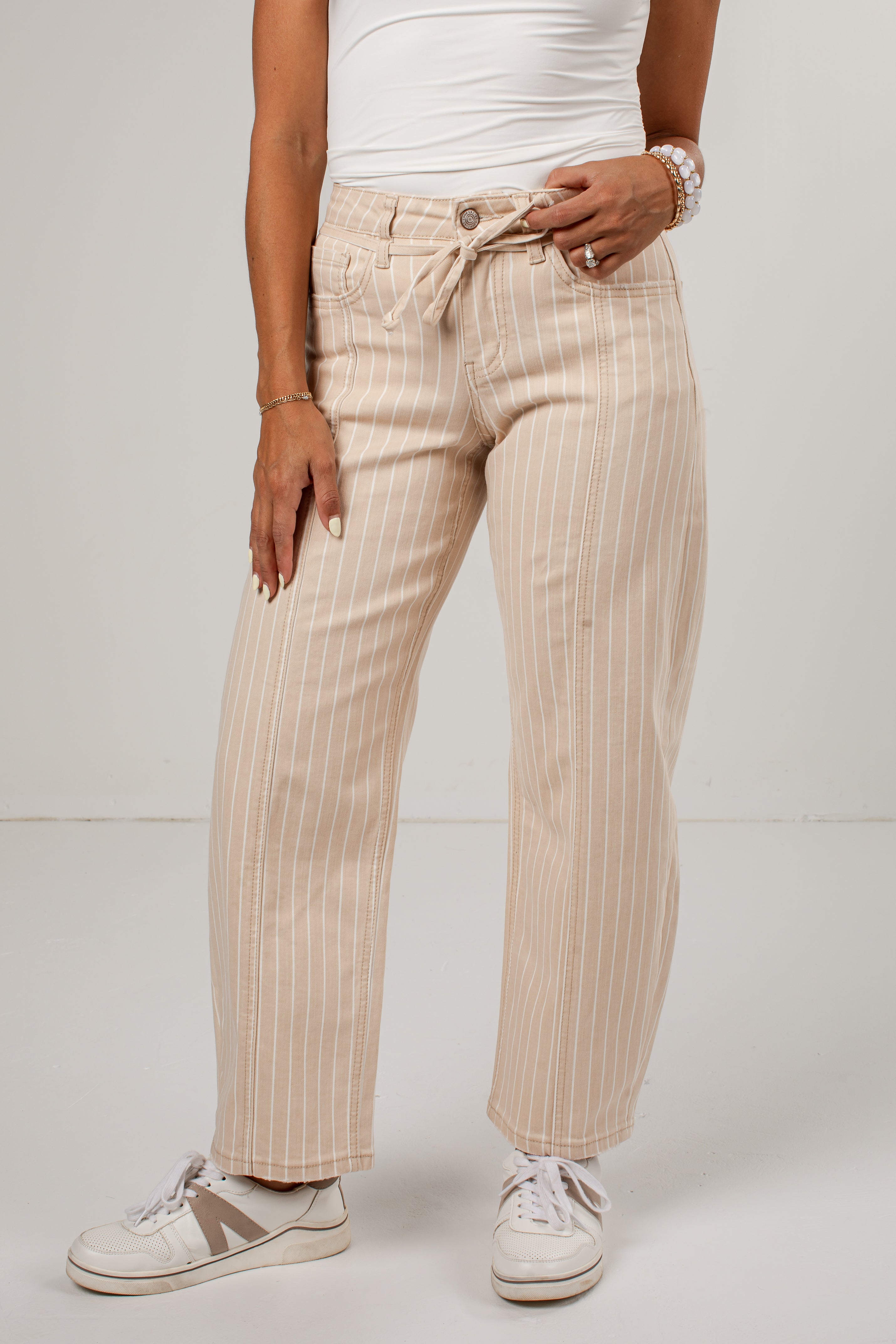 A person wearing beige pinstriped jeans with a drawstring waist, styled with a white tank top and white sneakers against a plain light gray studio background.
