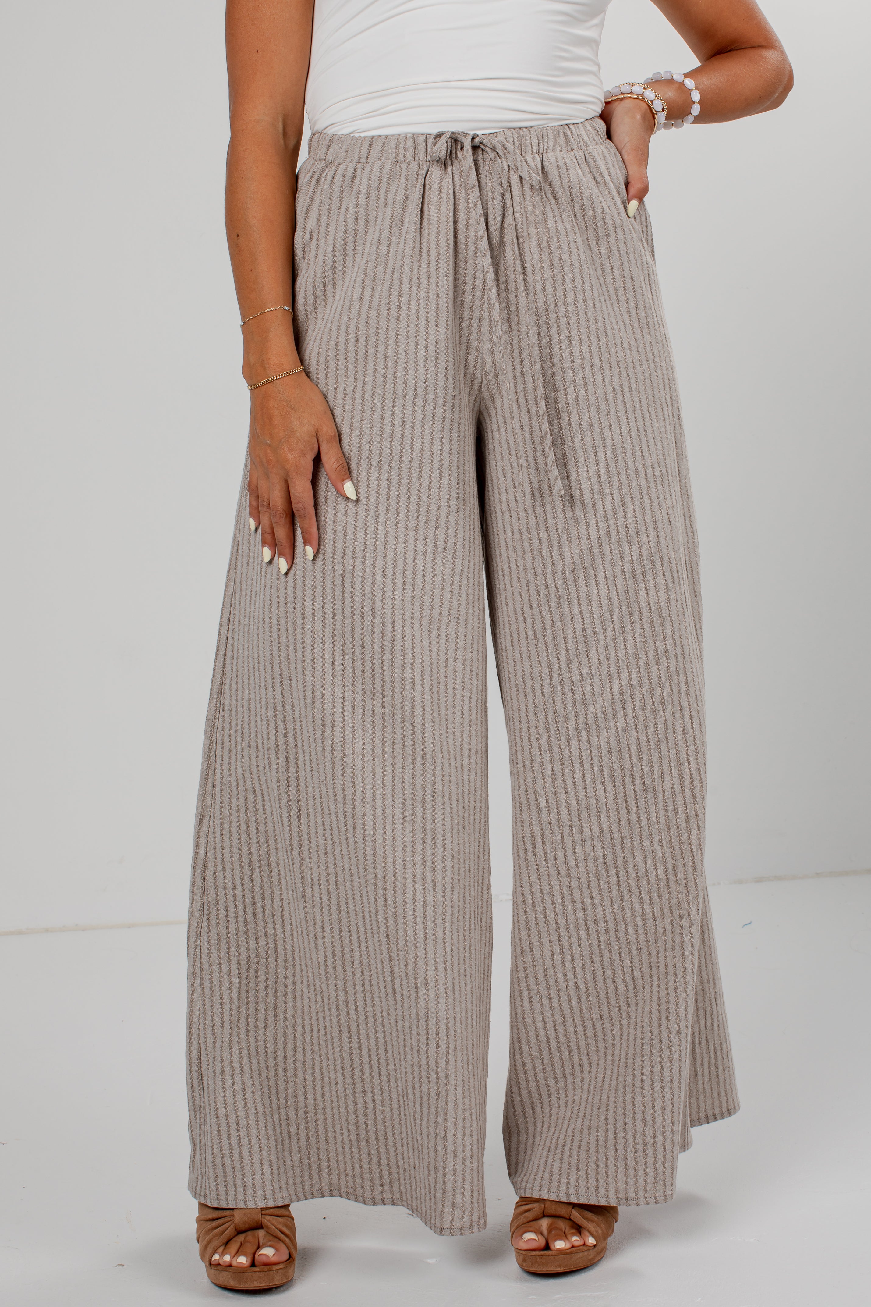A person wearing wide-leg pleated pants in a taupe-beige color with thin vertical stripes, paired with a white top and brown sandals, shown from the waist down against a light gray studio background.