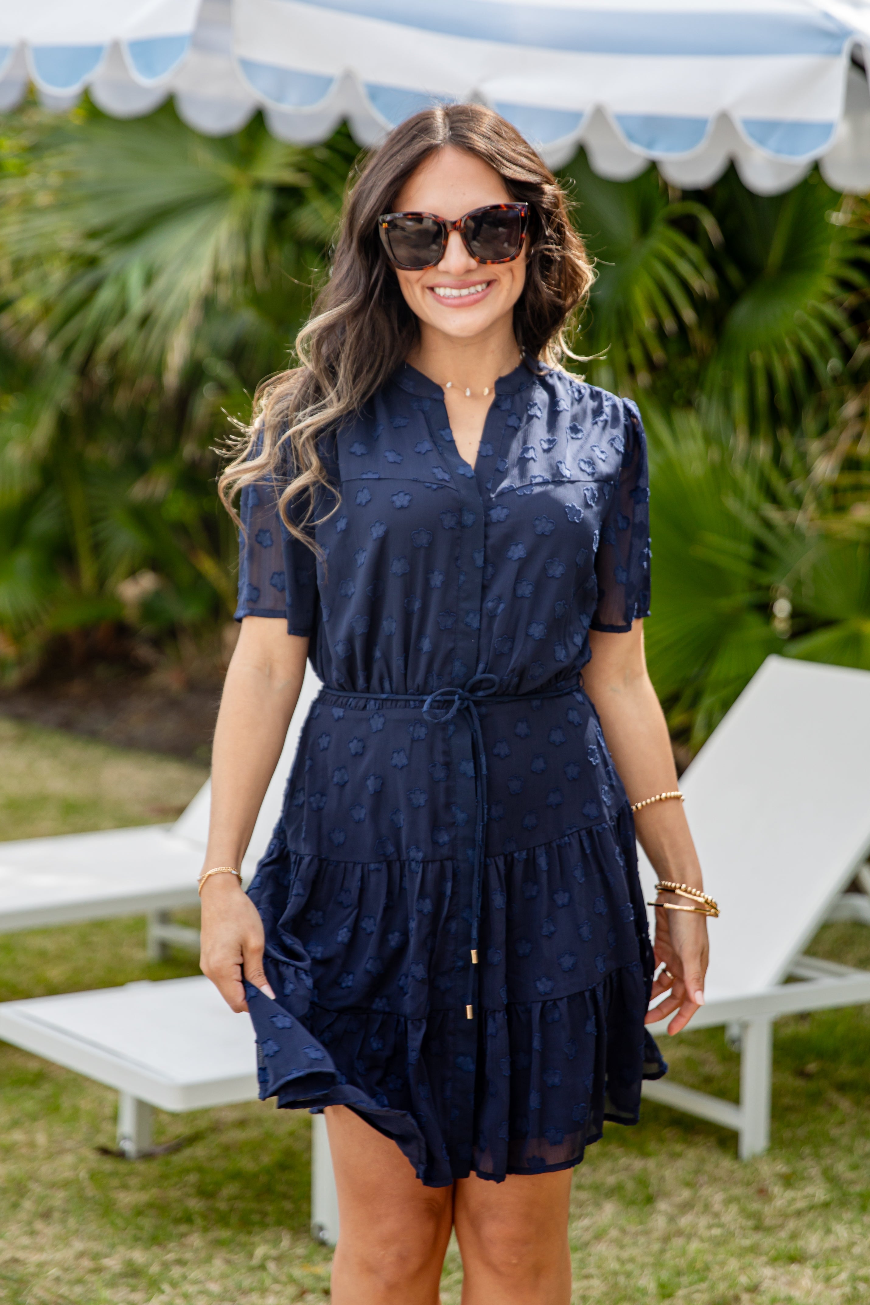 Woman wearing a navy dress and sunglasses outdoors with greenery and white canopies in the background