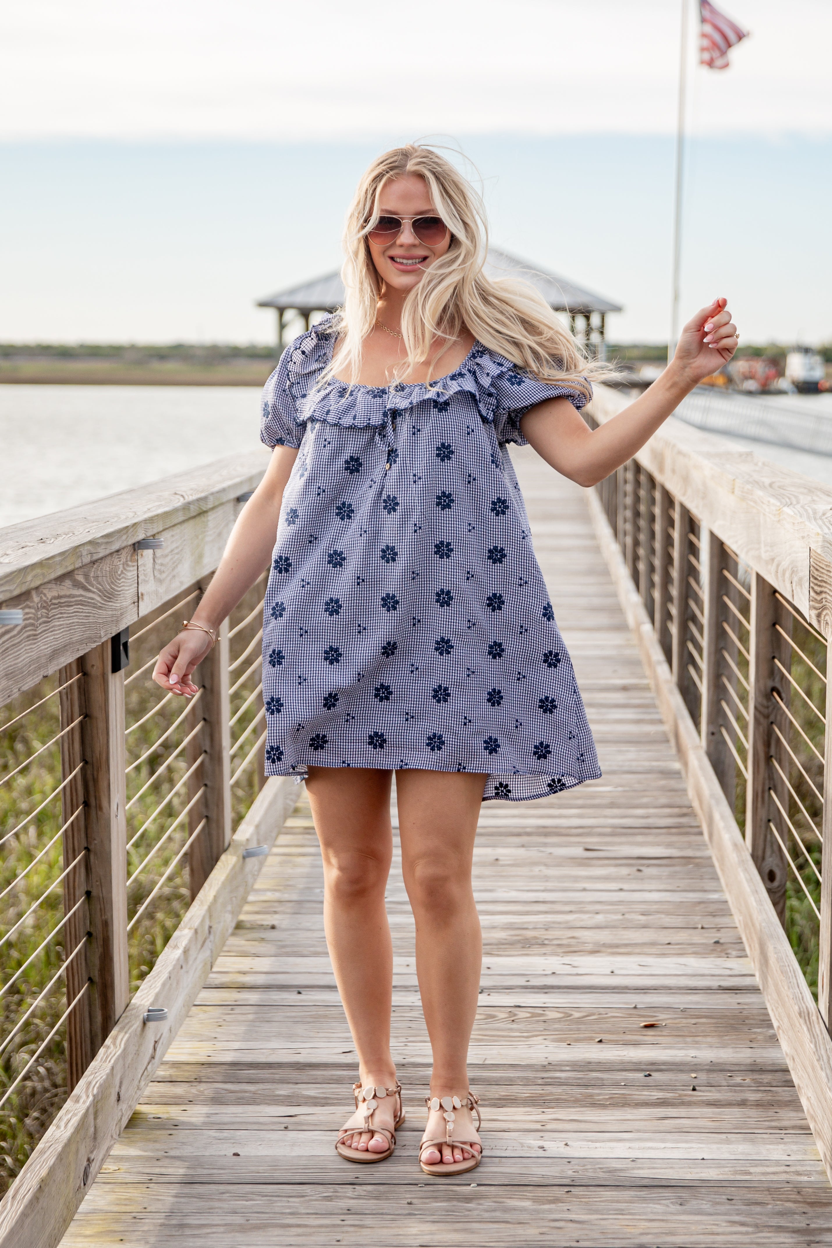 Woman in a blue floral dress standing on a wooden boardwalk by a waterfront.