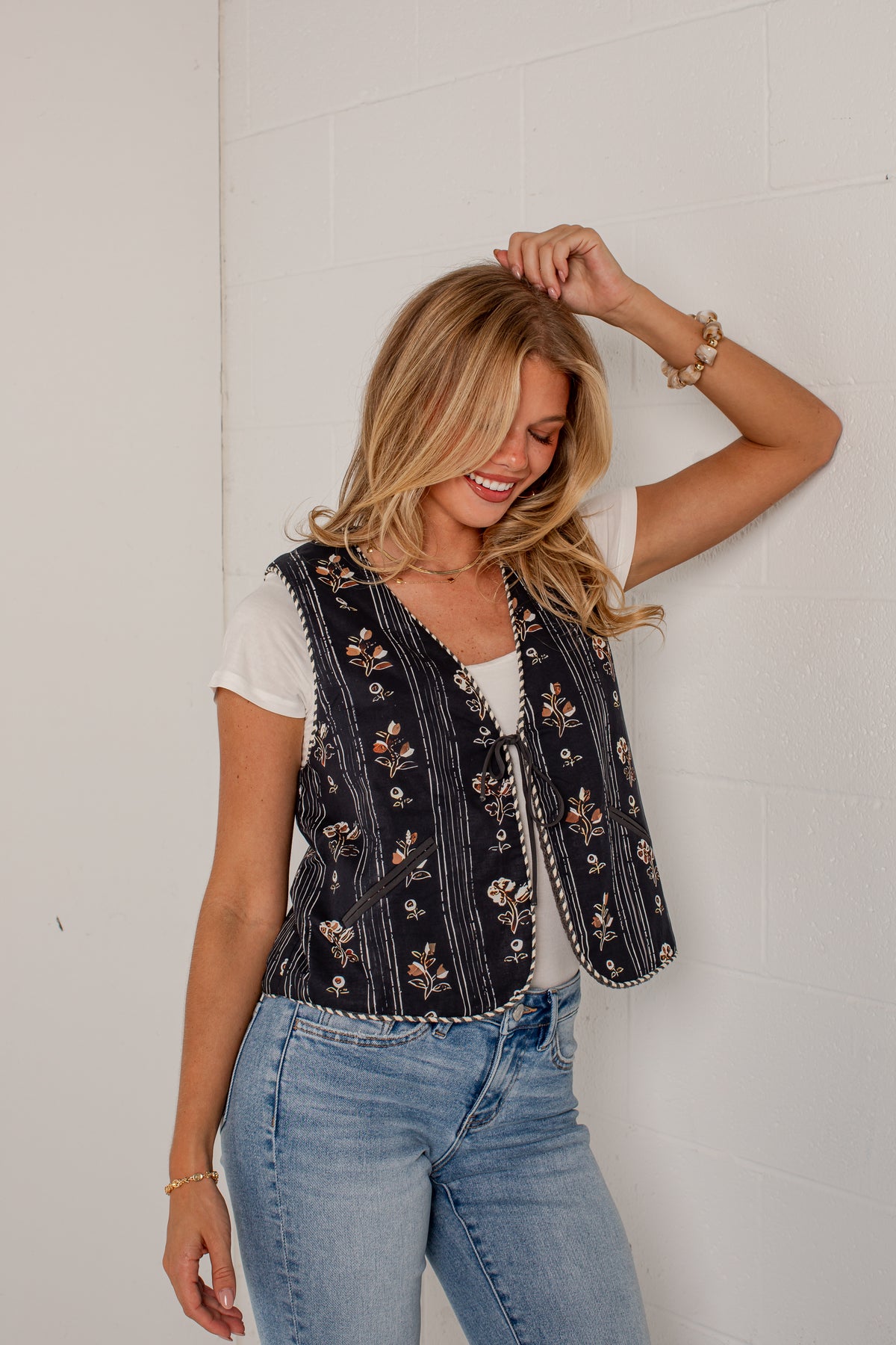 A woman wearing a sleeveless black vest with vertical white stripes and a floral pattern, styled over a white t-shirt and blue jeans, posing against a white brick wall.