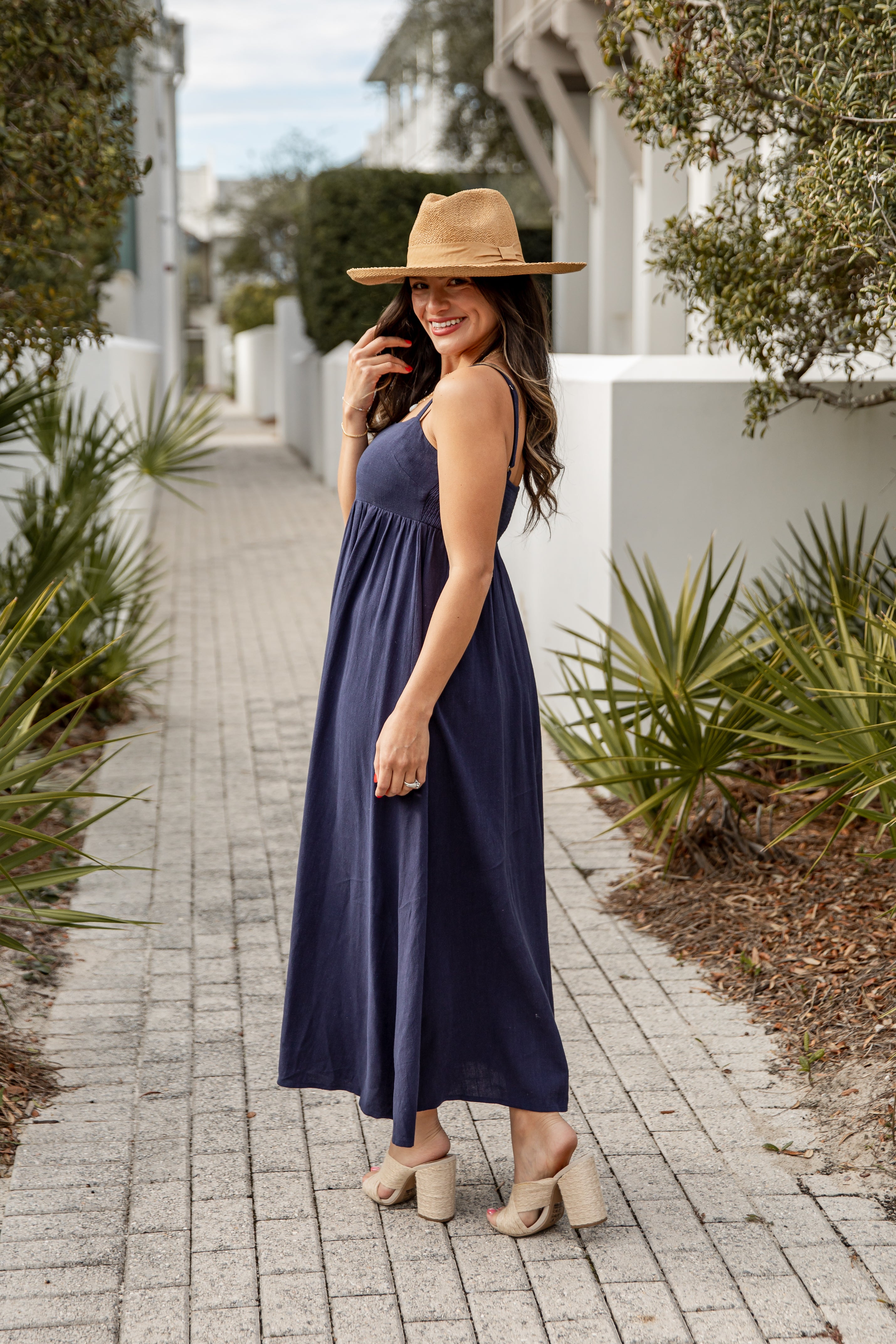 Woman in a navy dress and beige hat walking on a sidewalk with plants and a building in the background